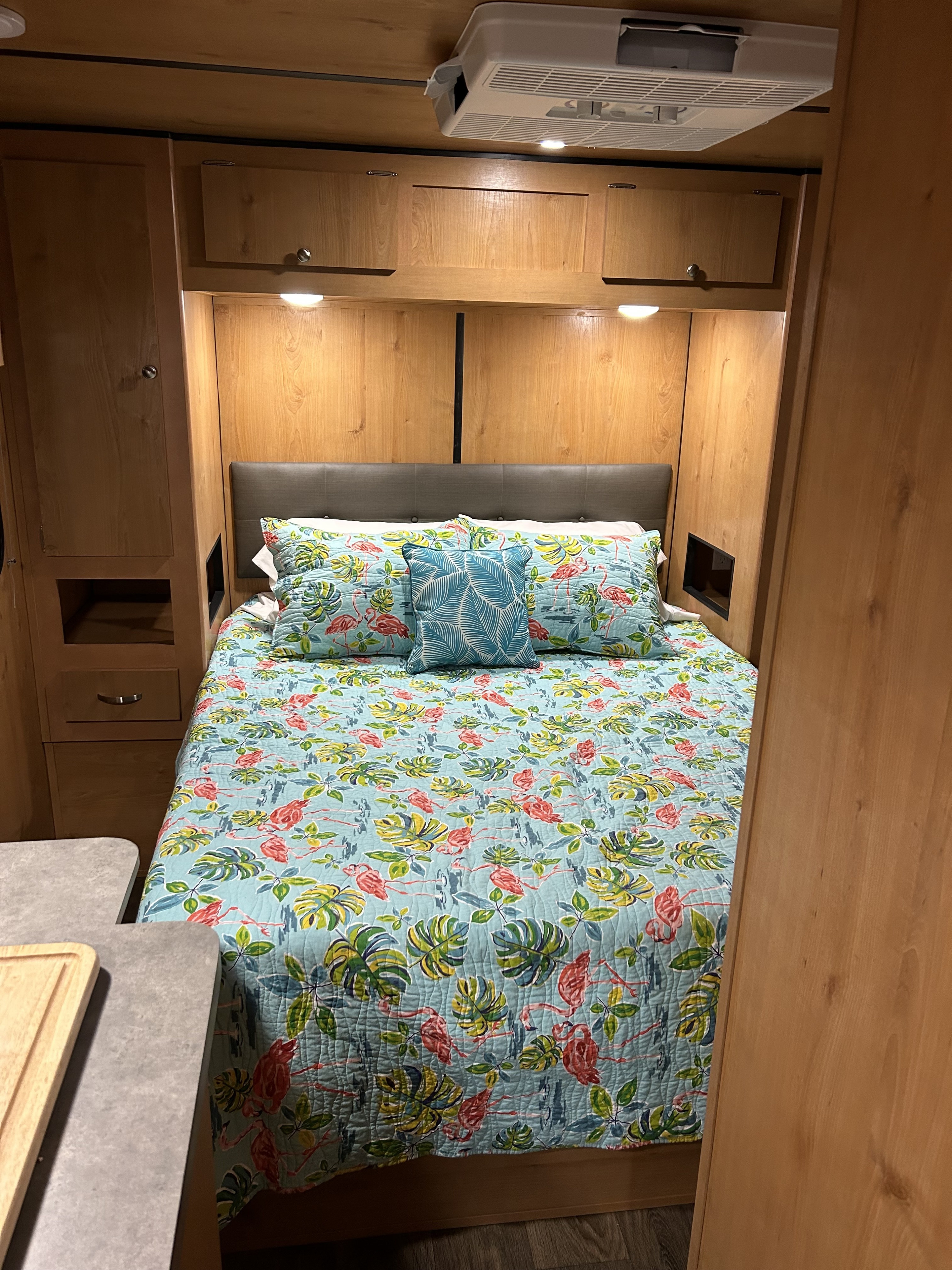 Cozy RV rental bedroom featuring a bed with vibrant flamingo and palm leaf bedding, overhead cabinets, and an AC unit.. Old School Trailer Other 2023