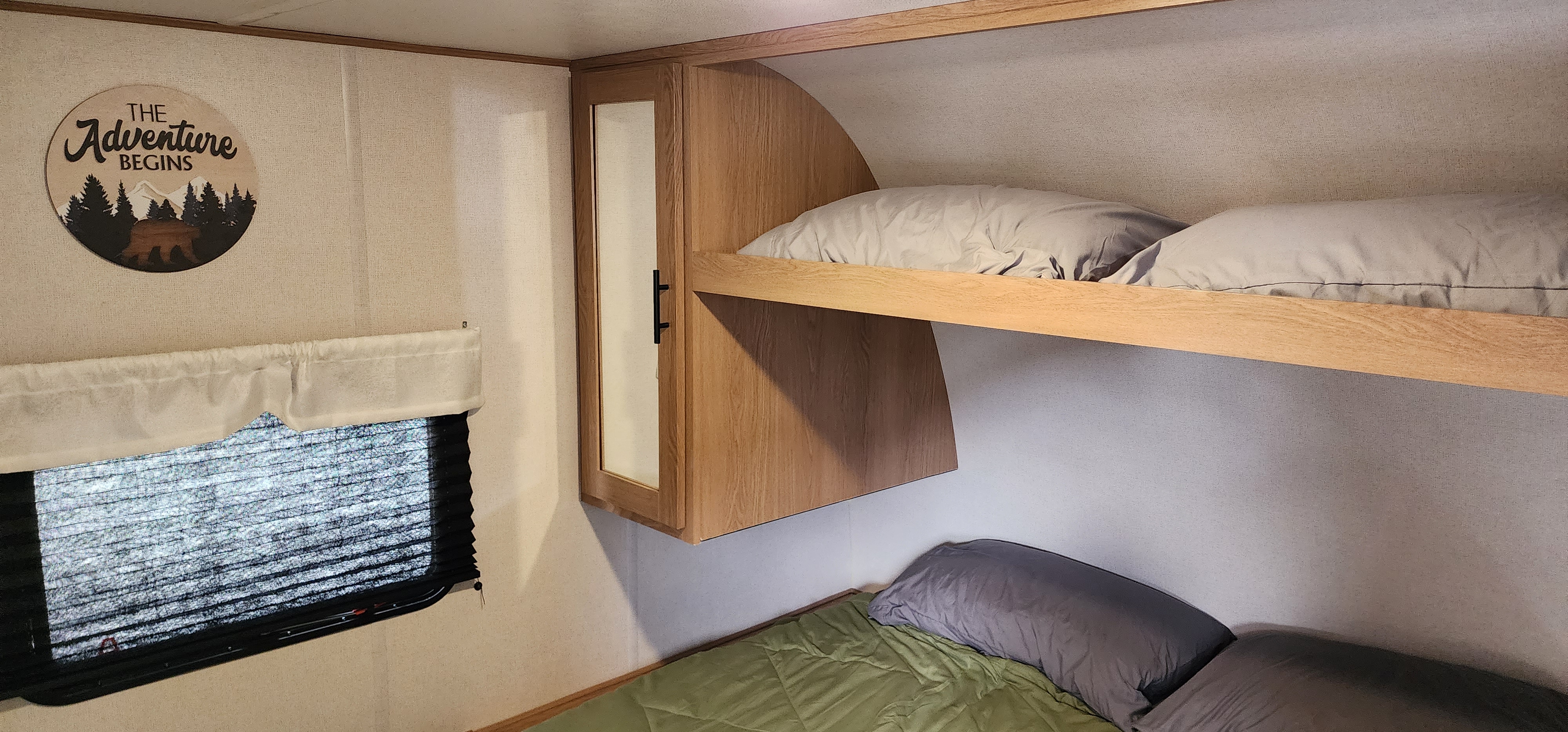 This RV rental features two stacked light wood bunk beds with bedding, a mirrored cabinet, and a window. A 'The Adventure Begins' sign is on the wall.. Forest River Avenger 26DBSLE 2022