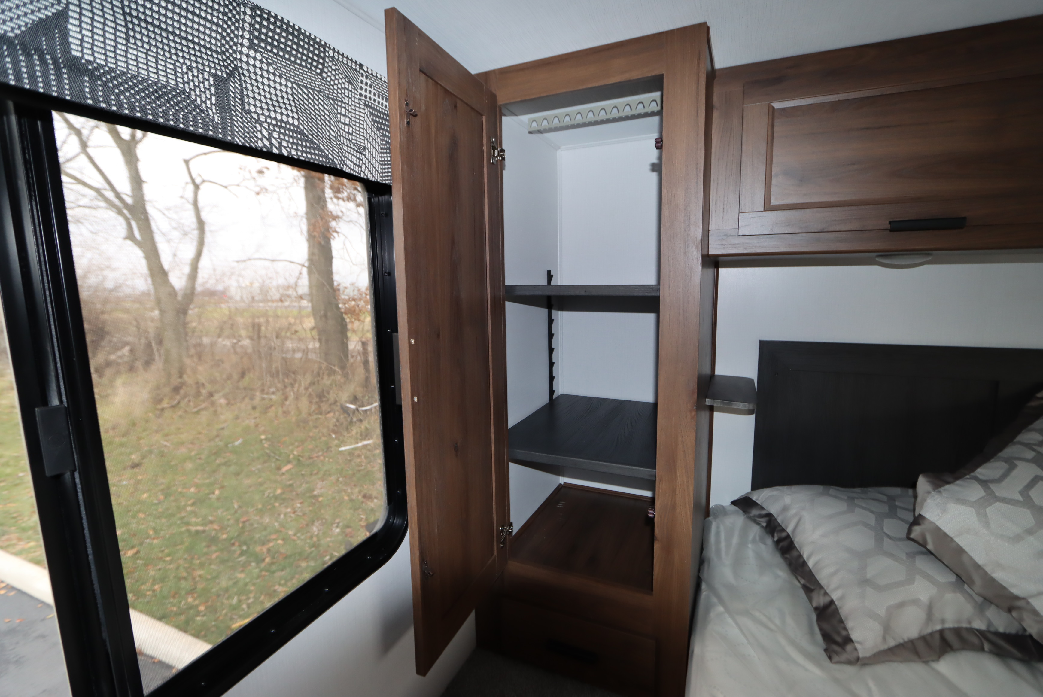 An RV rental bedroom offers a large window, an open cabinet with adjustable shelves, and overhead storage above a bed.. Forest River Sunseeker LE 2022