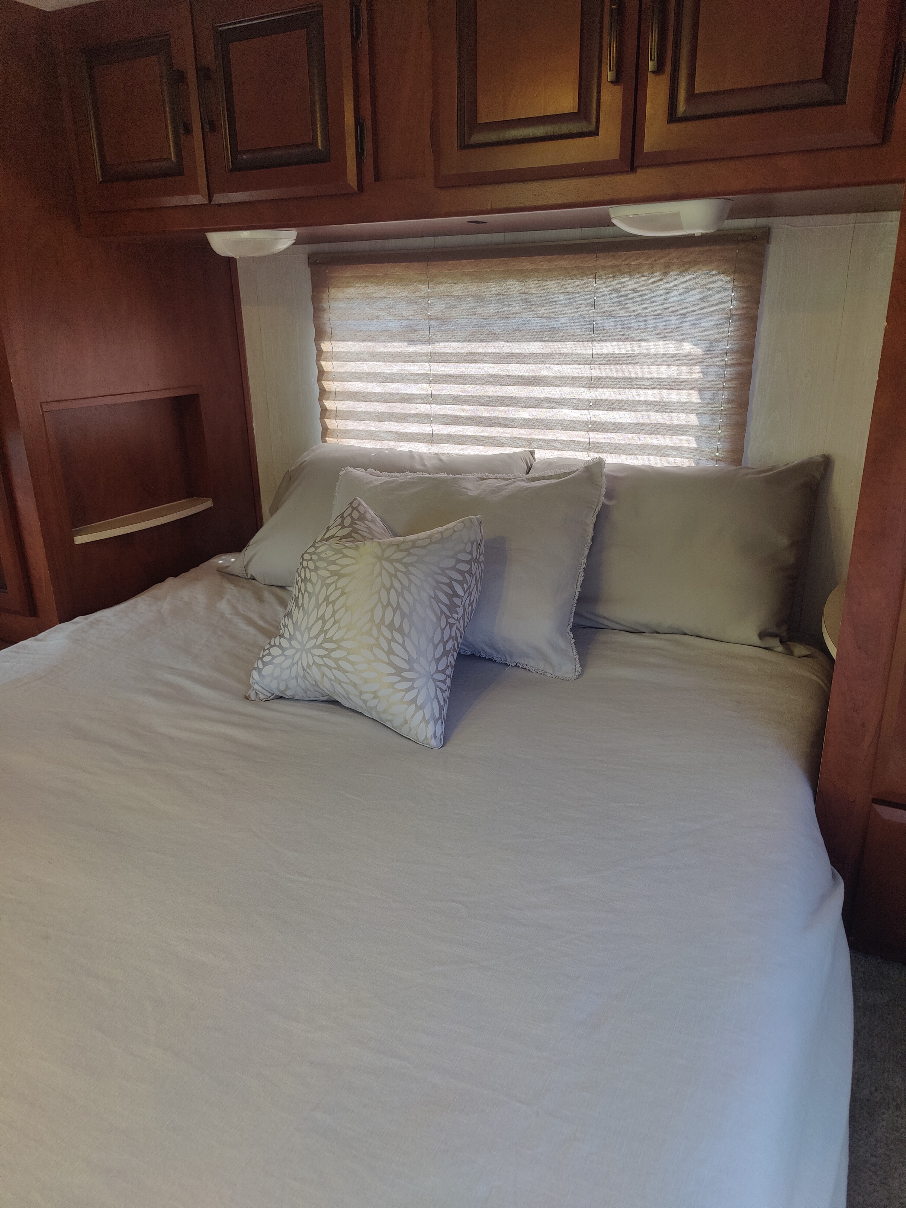 Comfortable RV rental bedroom featuring a made bed with pillows, overhead wooden cabinets, and a window with blinds.. Forest River Sunseeker 2012