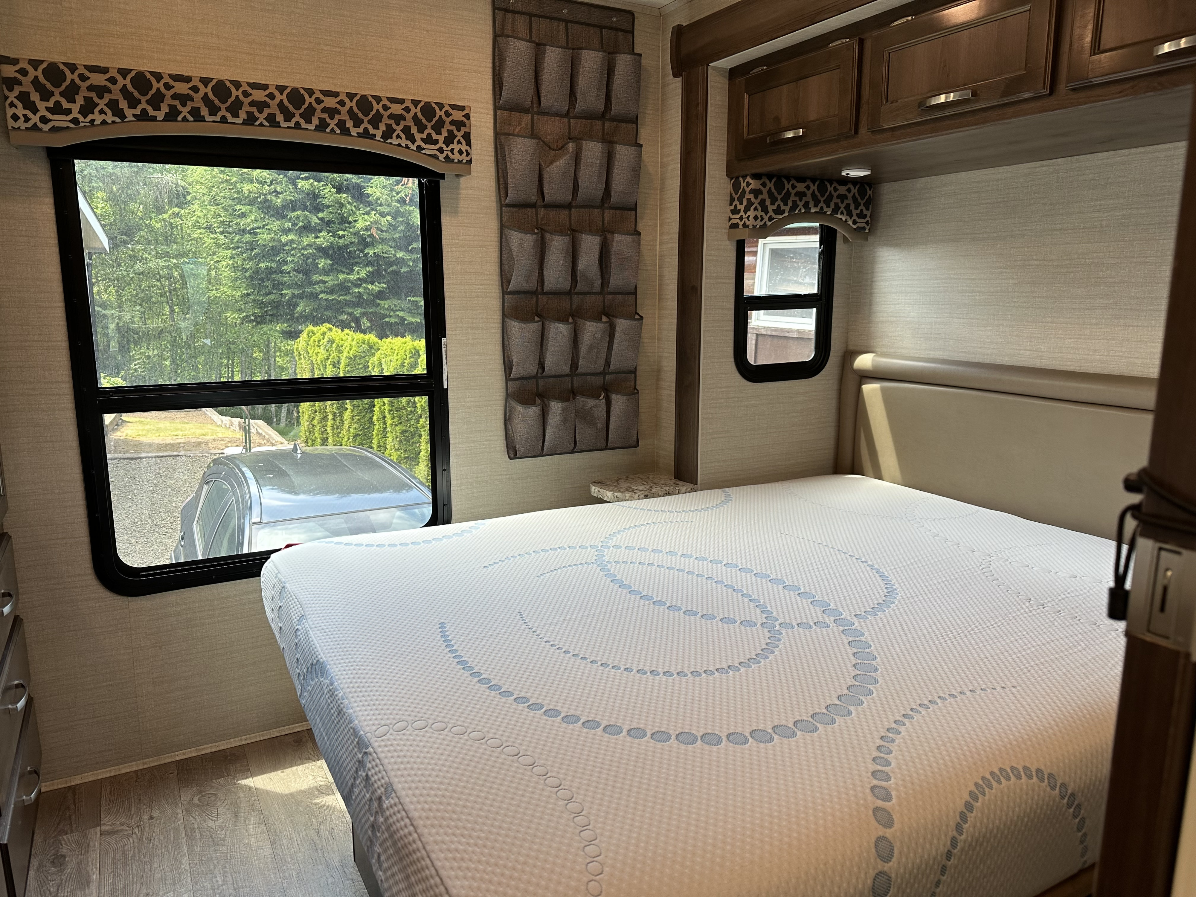 Inside an RV rental bedroom showing a bed, mattress with blue patterns, a large window with outdoor view, and hanging wall storage.. Jayco Alante 2018