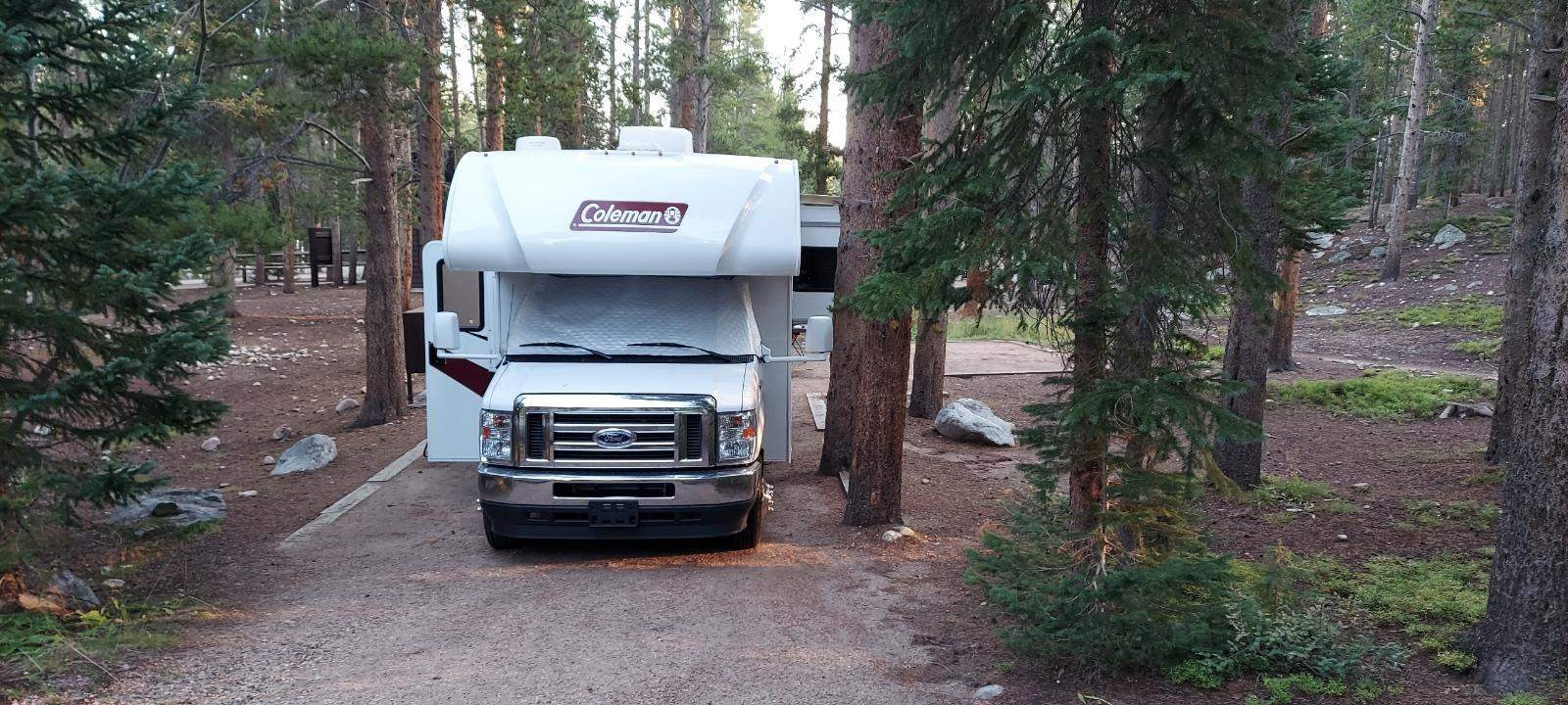White RV rental parked at a serene forest campsite, surrounded by tall pine trees, ideal for outdoor adventures and nature trips.. Thor COLEMAN 2023