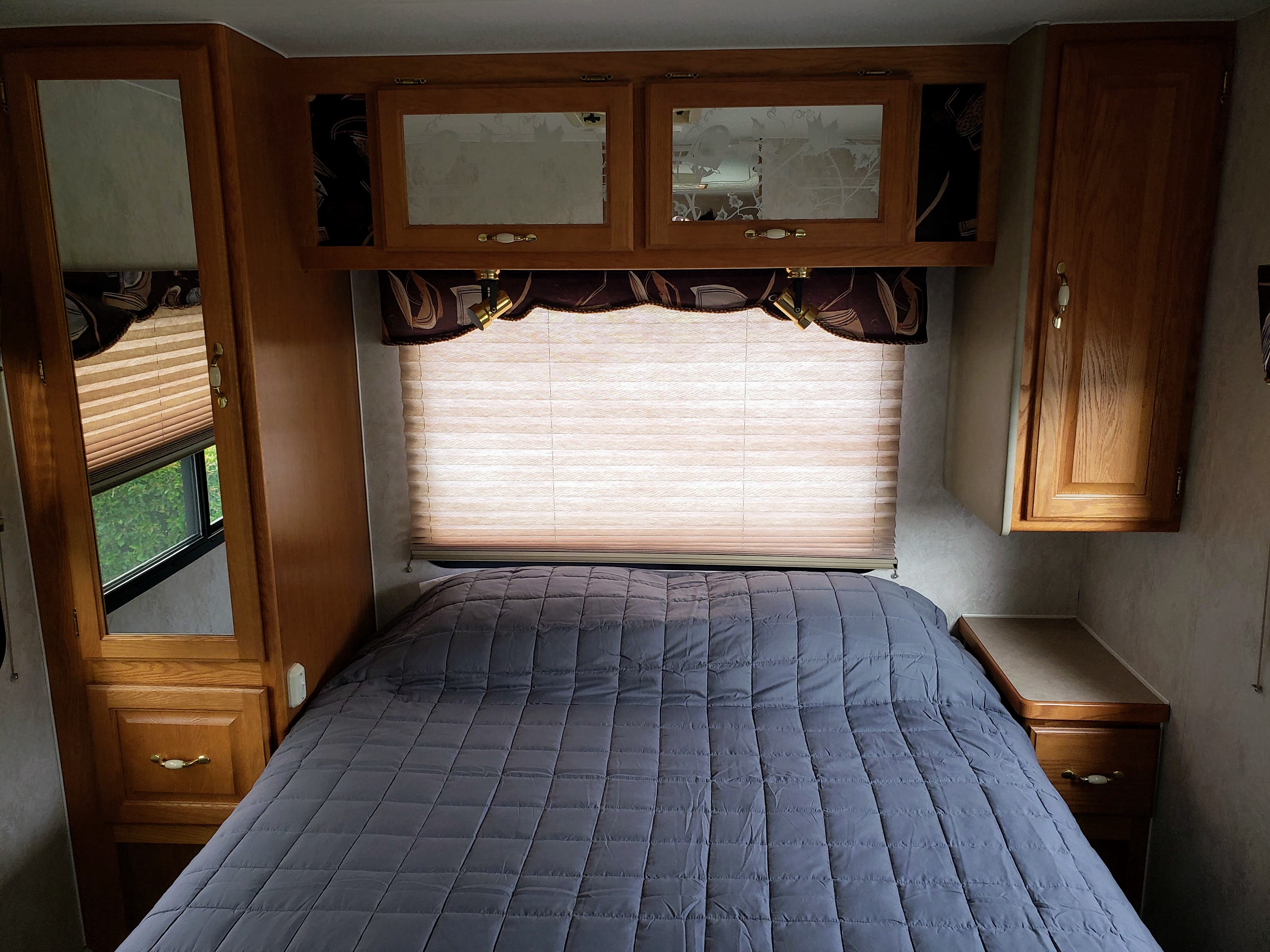 Cozy RV rental bedroom featuring a gray quilted bed, a window with a blind, and ample wood cabinetry.. Triple E Regency 1999