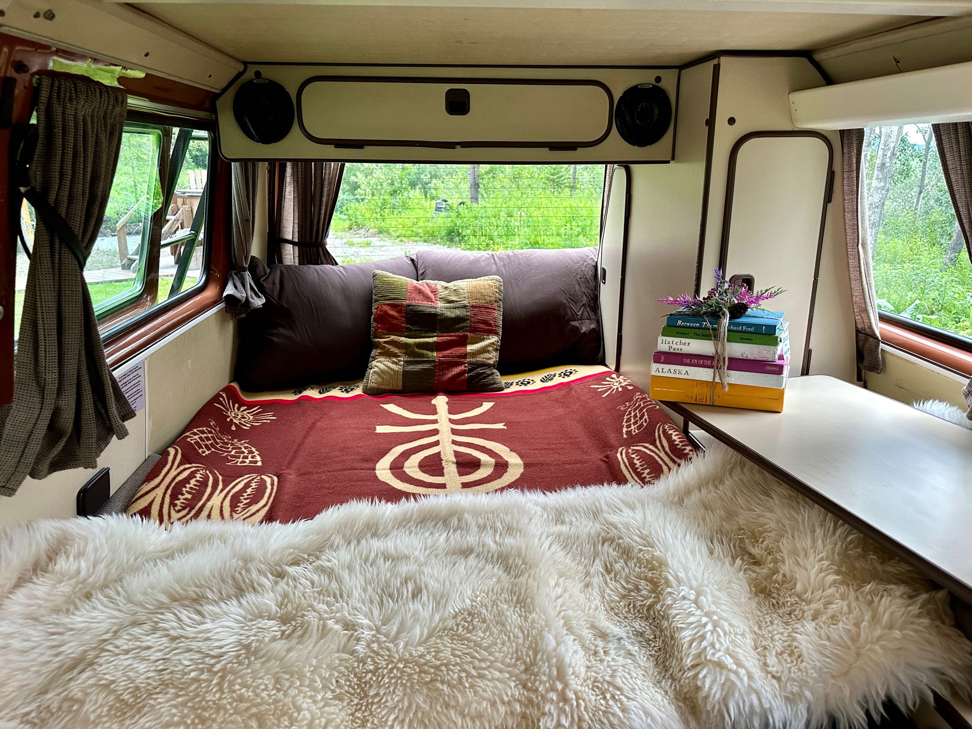 RV rental interior featuring a cozy bed with a furry blanket, a stack of books, and a beautiful green nature view.. Volkswagen Westfalia 1984