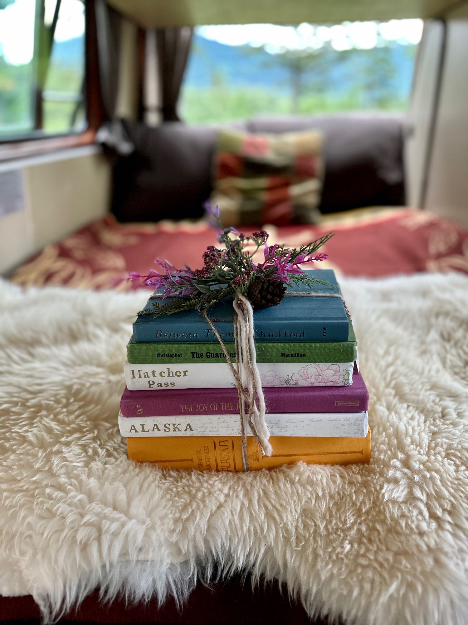 Cozy RV rental: A stack of colorful travel books, adorned with flowers and twine, rests on a fluffy white rug, with a blurred mountain view outside.. Volkswagen Westfalia 1984