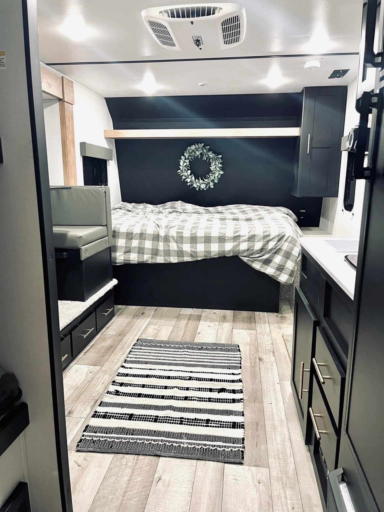 Modern RV rental interior with a bed, bench seating, and kitchenette. Features dark walls, plaid bedding, and light wood floors.. Keystone RV Springdale 2022