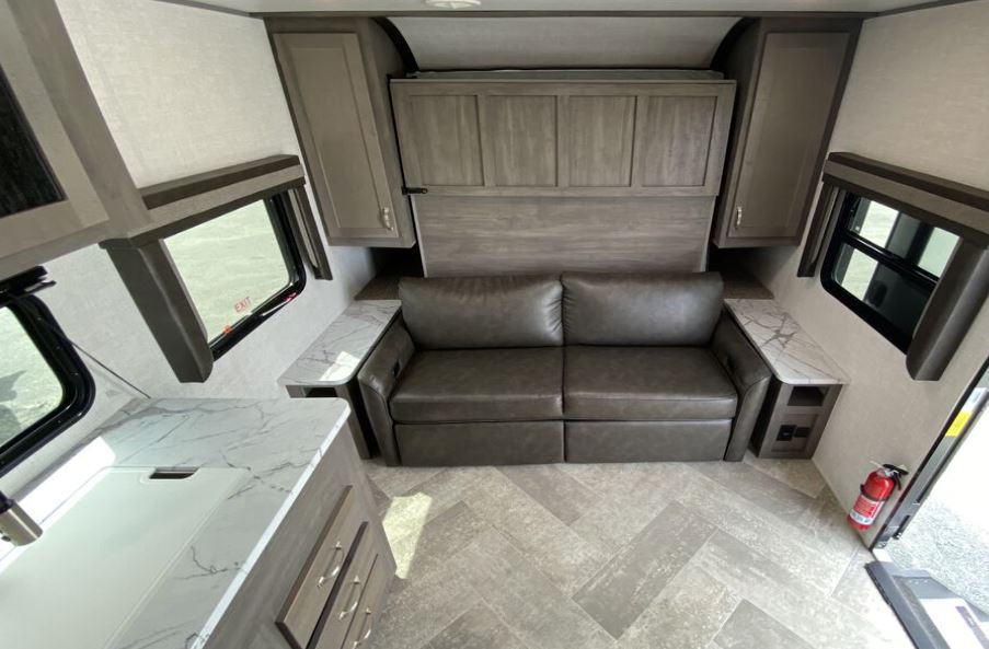 This RV rental interior shows a grey sofa, overhead cabinets, and a versatile bed system. A white countertop kitchen is on the left.. East to West Alta 2021