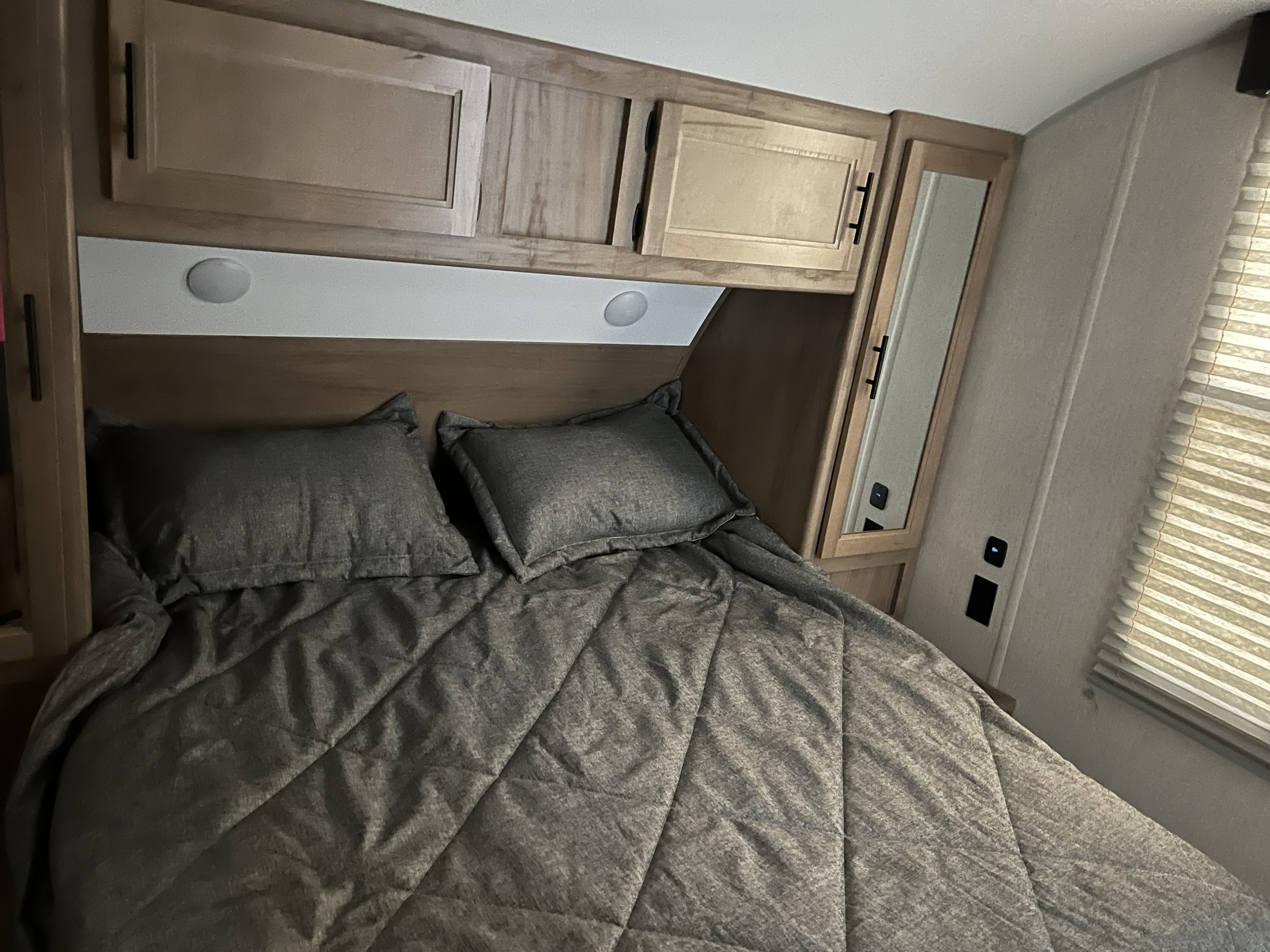 This RV rental bedroom offers a comfortable gray bed, plump pillows, overhead wood cabinets, and convenient bedside lighting.. Forest River 29TE 2023