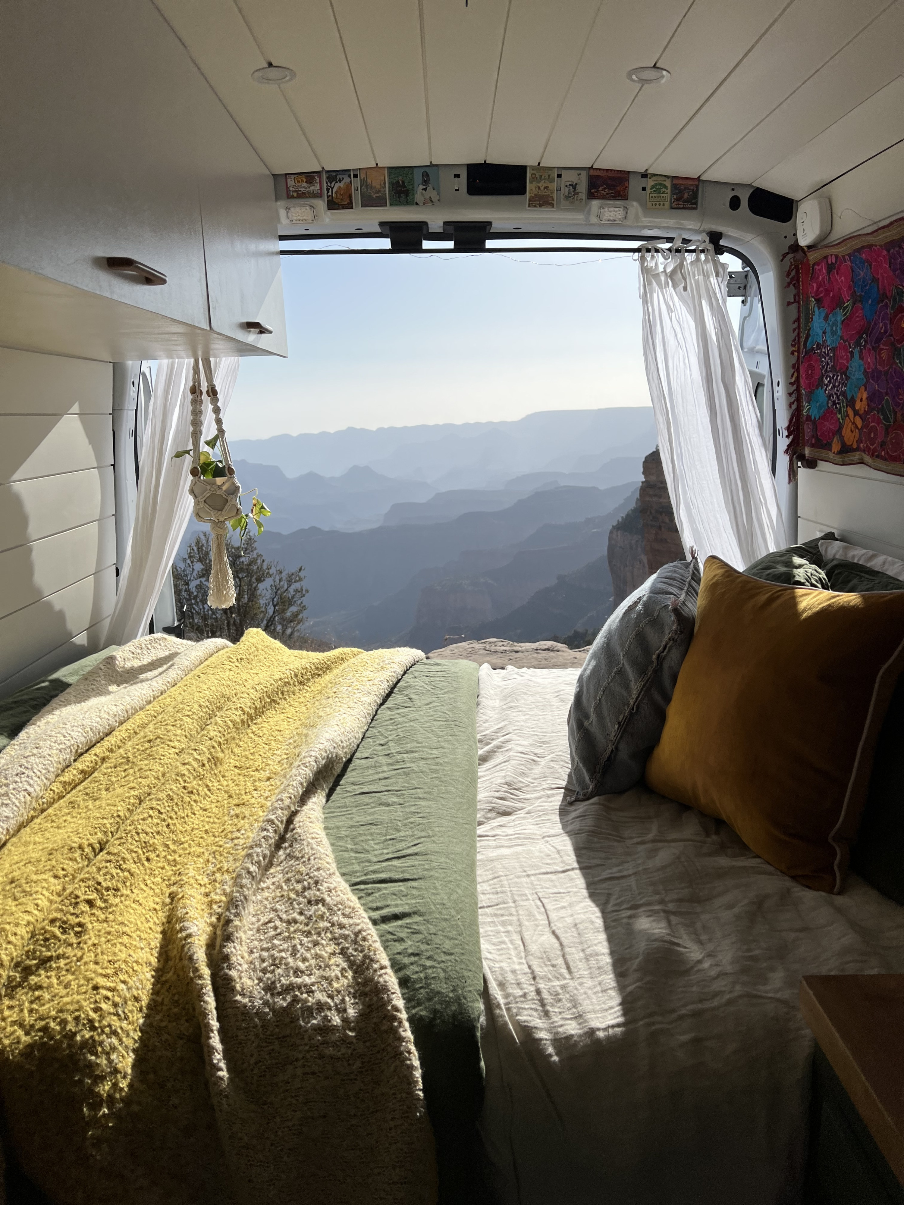 Inside a comfy RV rental, a bed with soft blankets faces an open back, revealing layers of serene mountain canyons.. Ford Transit 2020