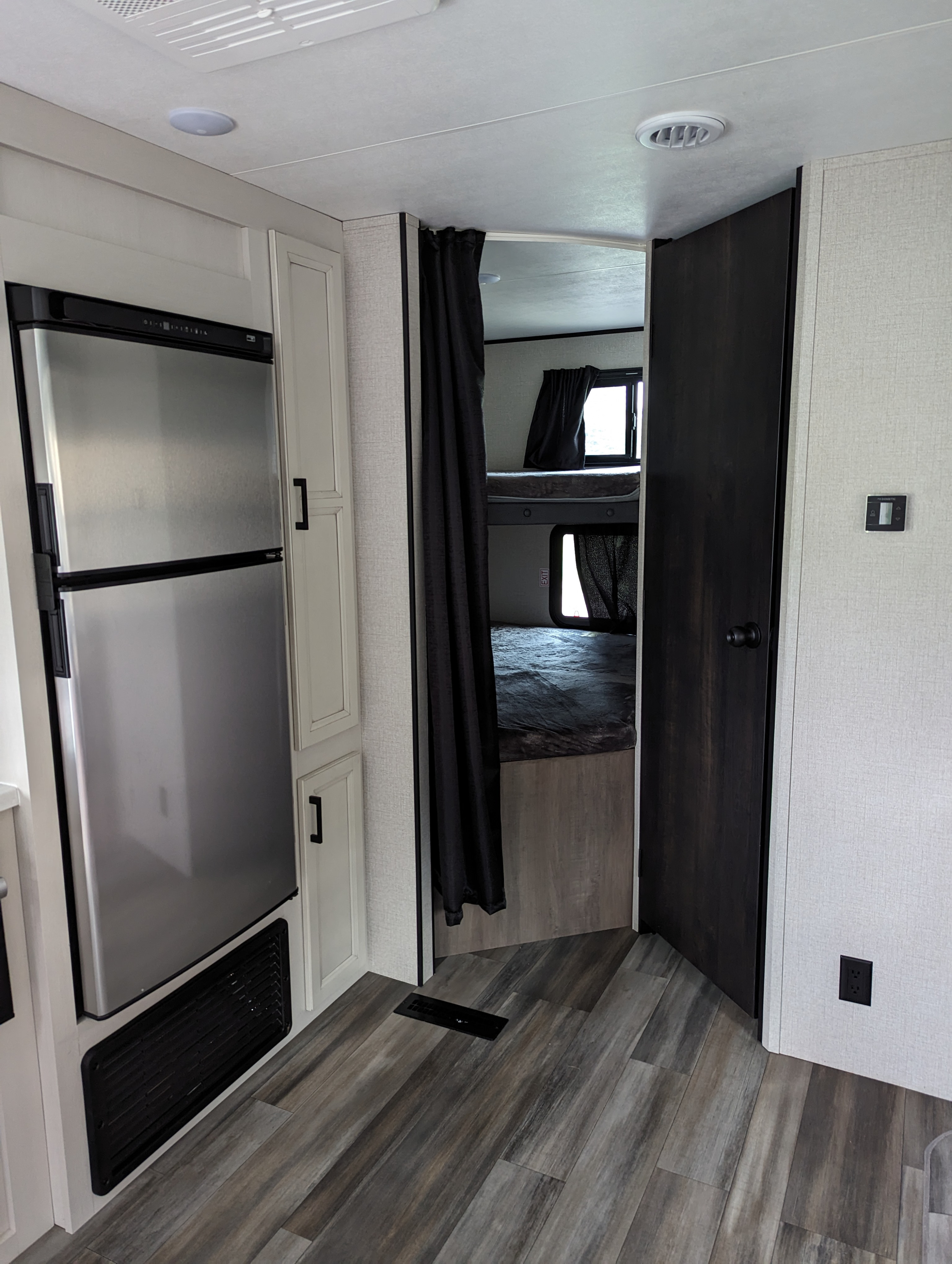A modern RV rental interior featuring a stainless steel fridge, light cabinets, and a bunk bed room with dark curtains.. Other Other 2023