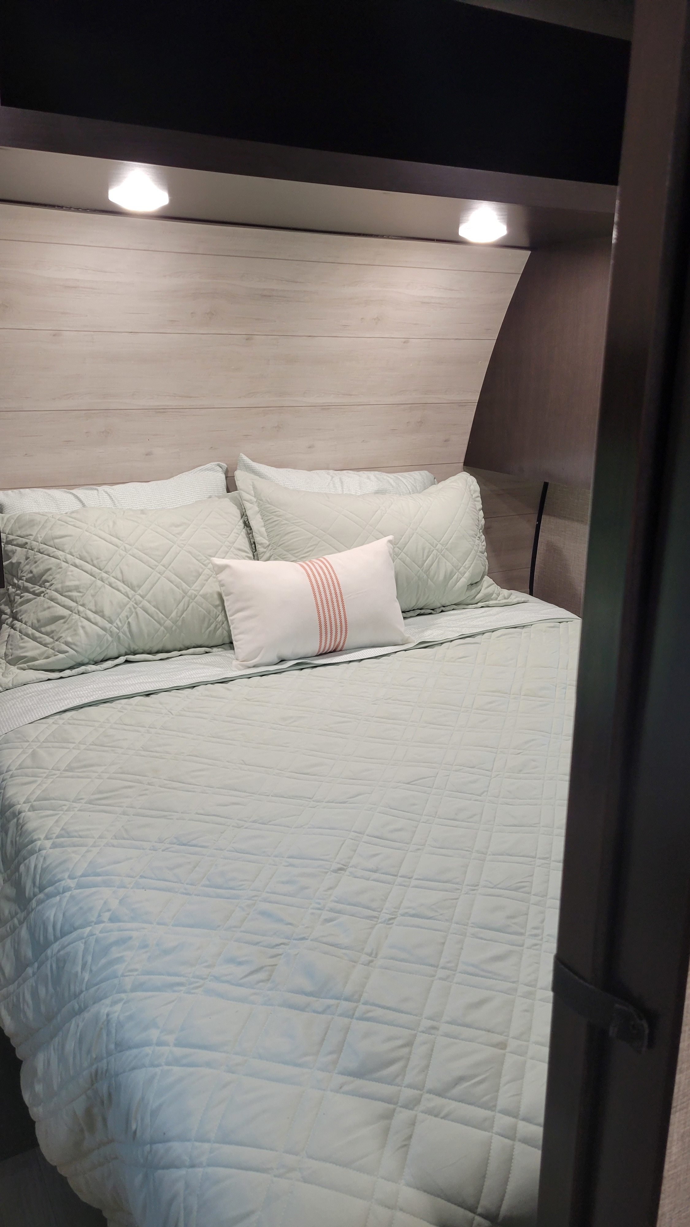 Cozy RV rental bedroom features a neatly made bed with light green quilted bedding, wood paneling, and bright overhead lights.. Jayco Jay Flight 2023