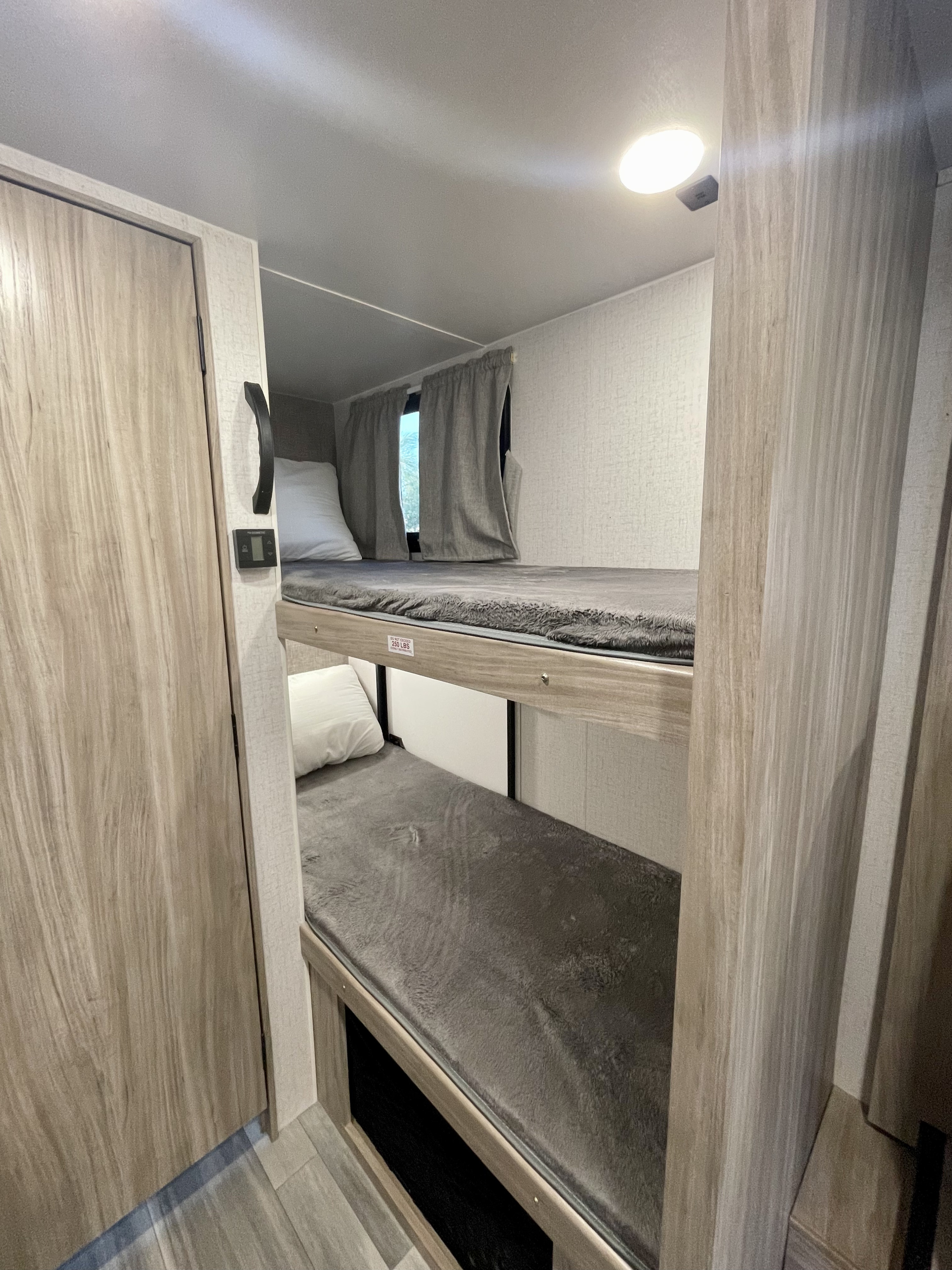 Inside an RV rental, two bunk beds with grey bedding and white pillows offer compact sleeping. A window with curtains and under-bed storage are visible.. Winnebago Micro Minnie 2022