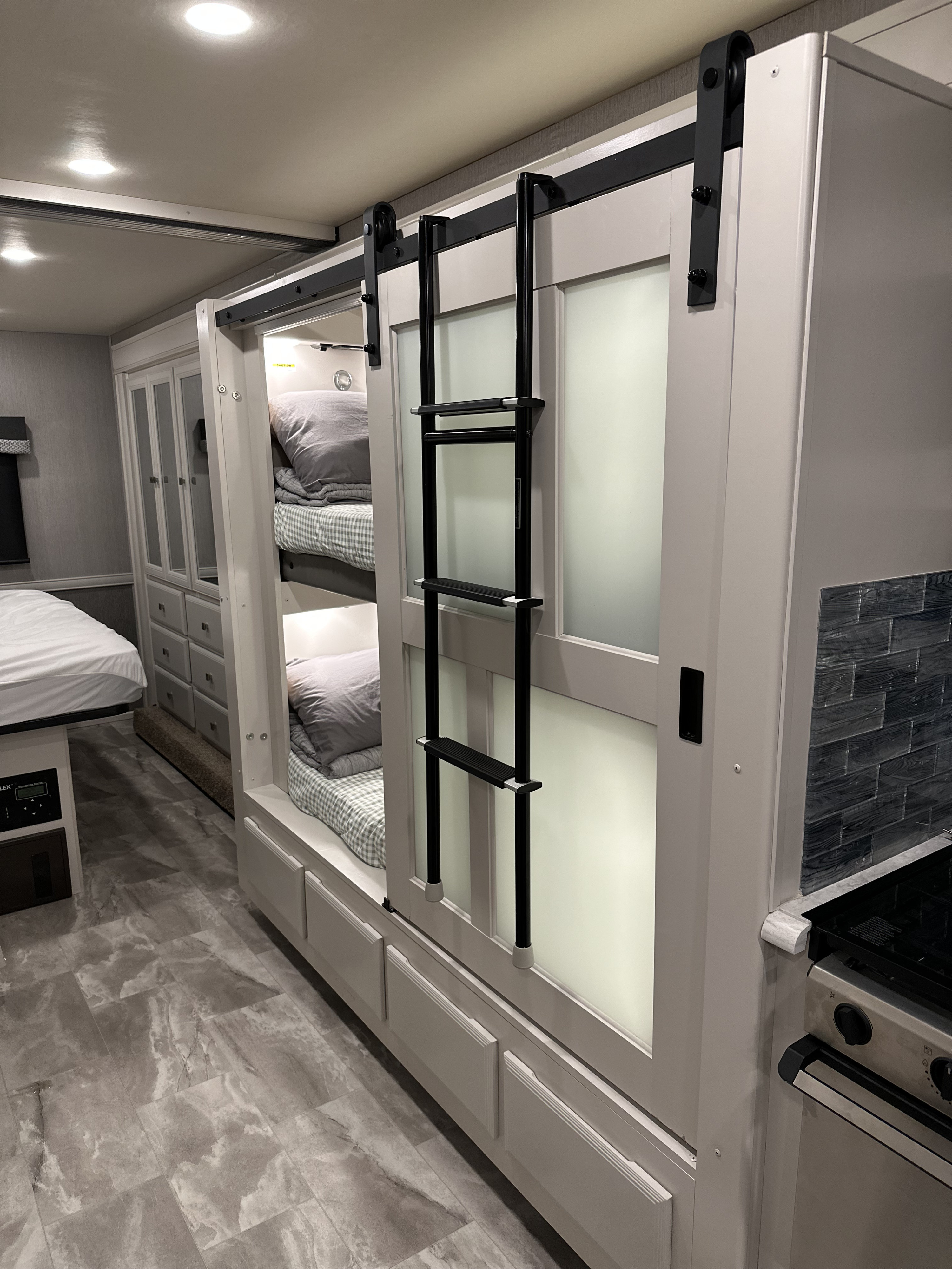 This RV rental interior showcases space-saving bunk beds with frosted sliding doors, a kitchen area, and a glimpse of a main bed.. Fleetwood Flair 2021