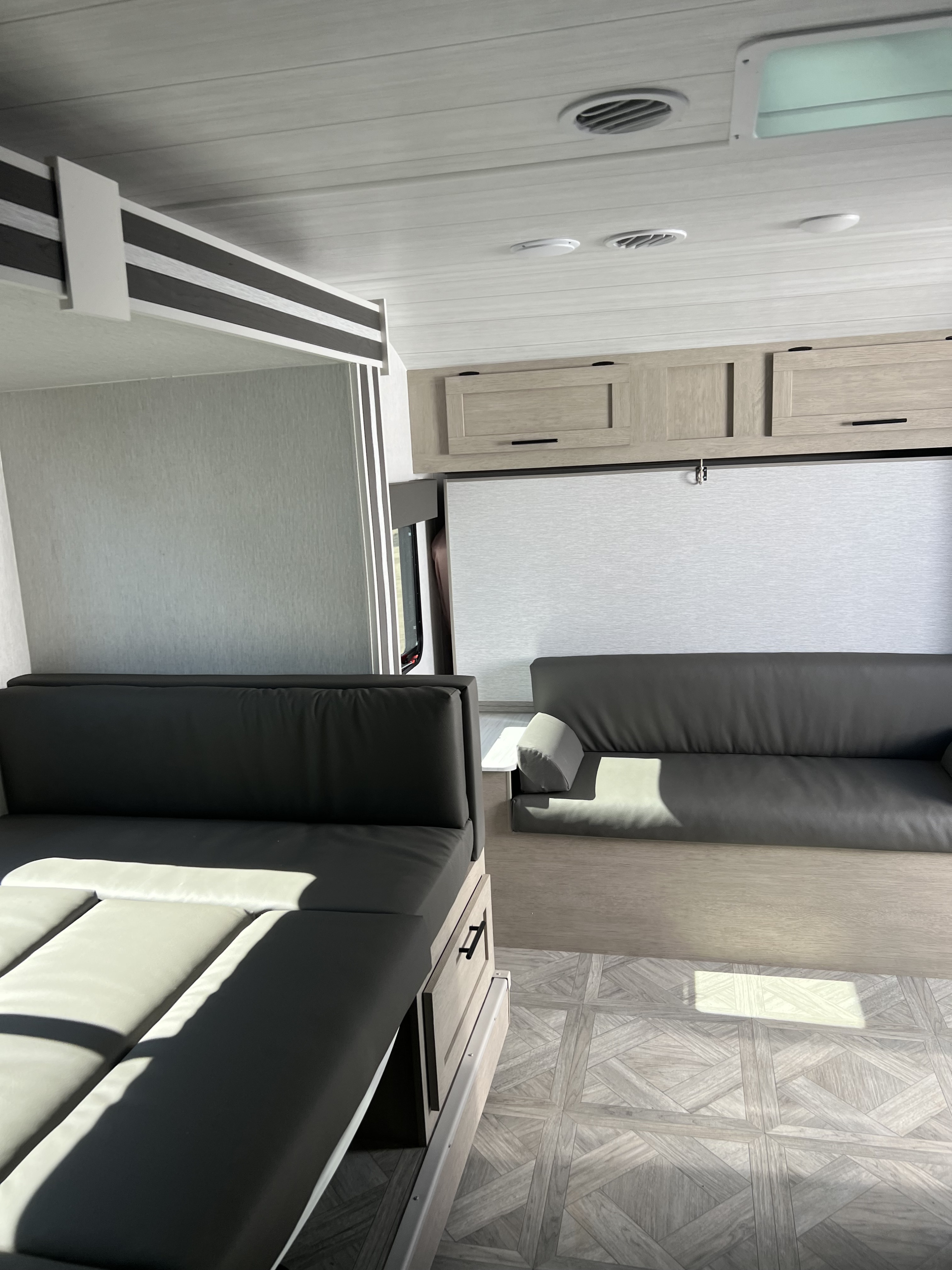 RV rental interior: two dark gray sofas, overhead cabinets, light wood patterned floor, and a sunlit window.. Forest River Wildwood FSX 2023