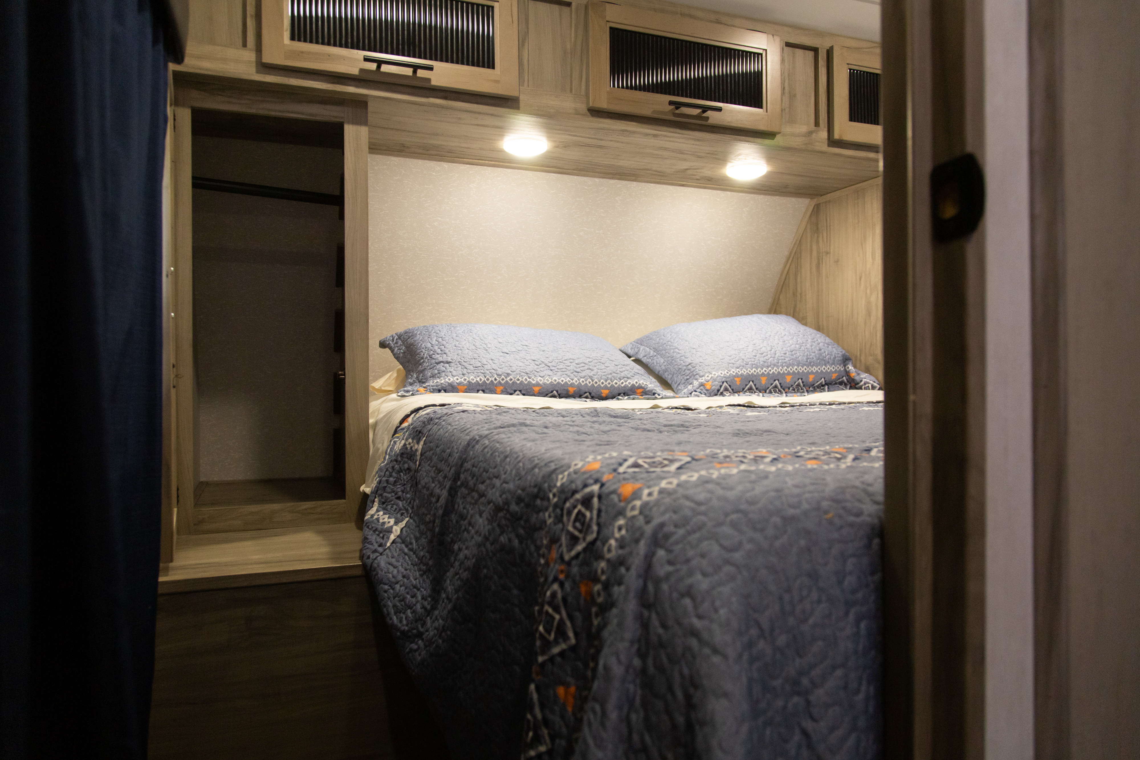 Step into comfort: This RV rental bedroom features a comfy bed with patterned bedding, overhead cabinets, and an open closet.. Forest River Alpha Wolf 2022