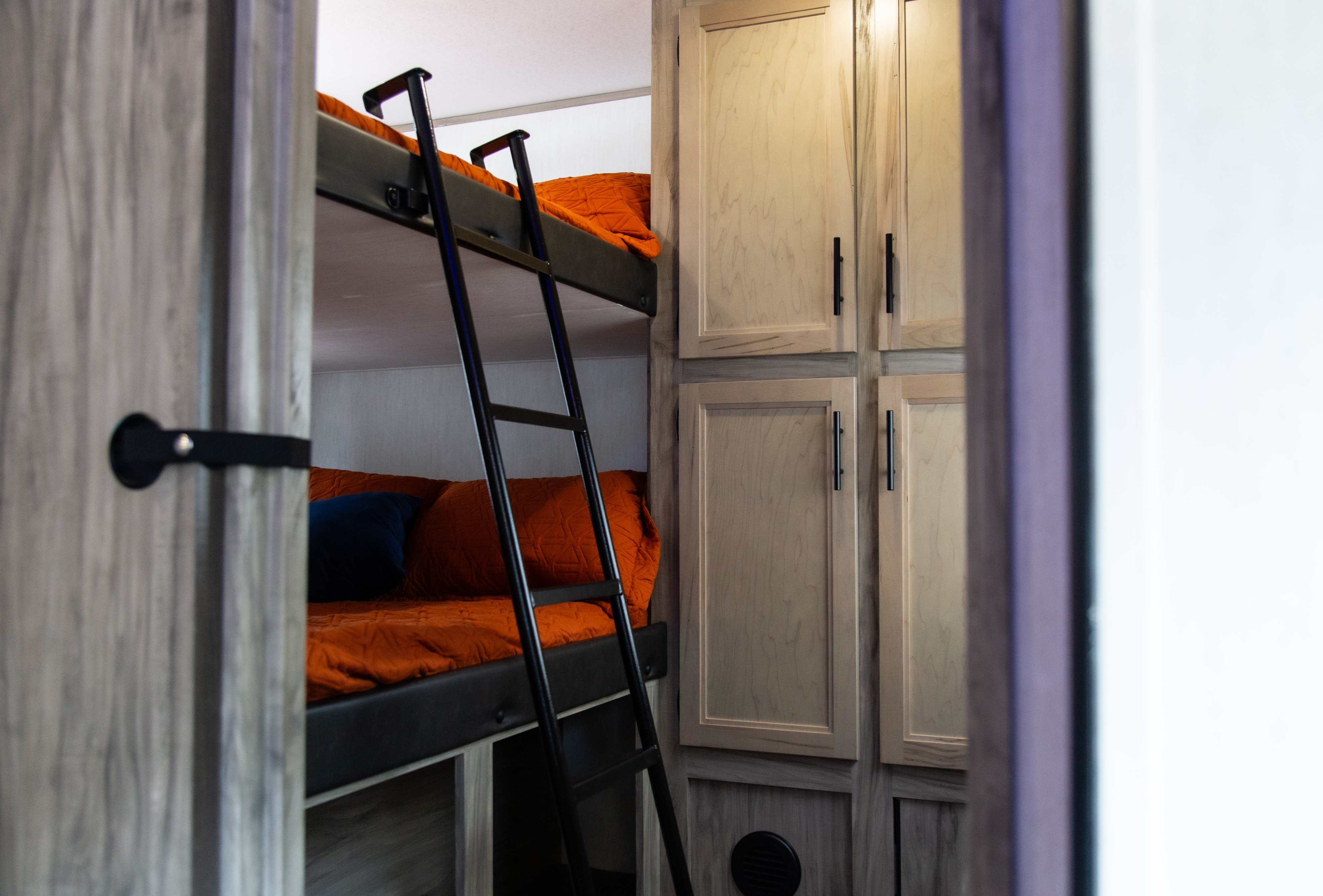 View into an RV rental bunk bed area featuring a ladder, vibrant orange bedding, and ample storage cabinets.. Forest River Alpha Wolf 2022