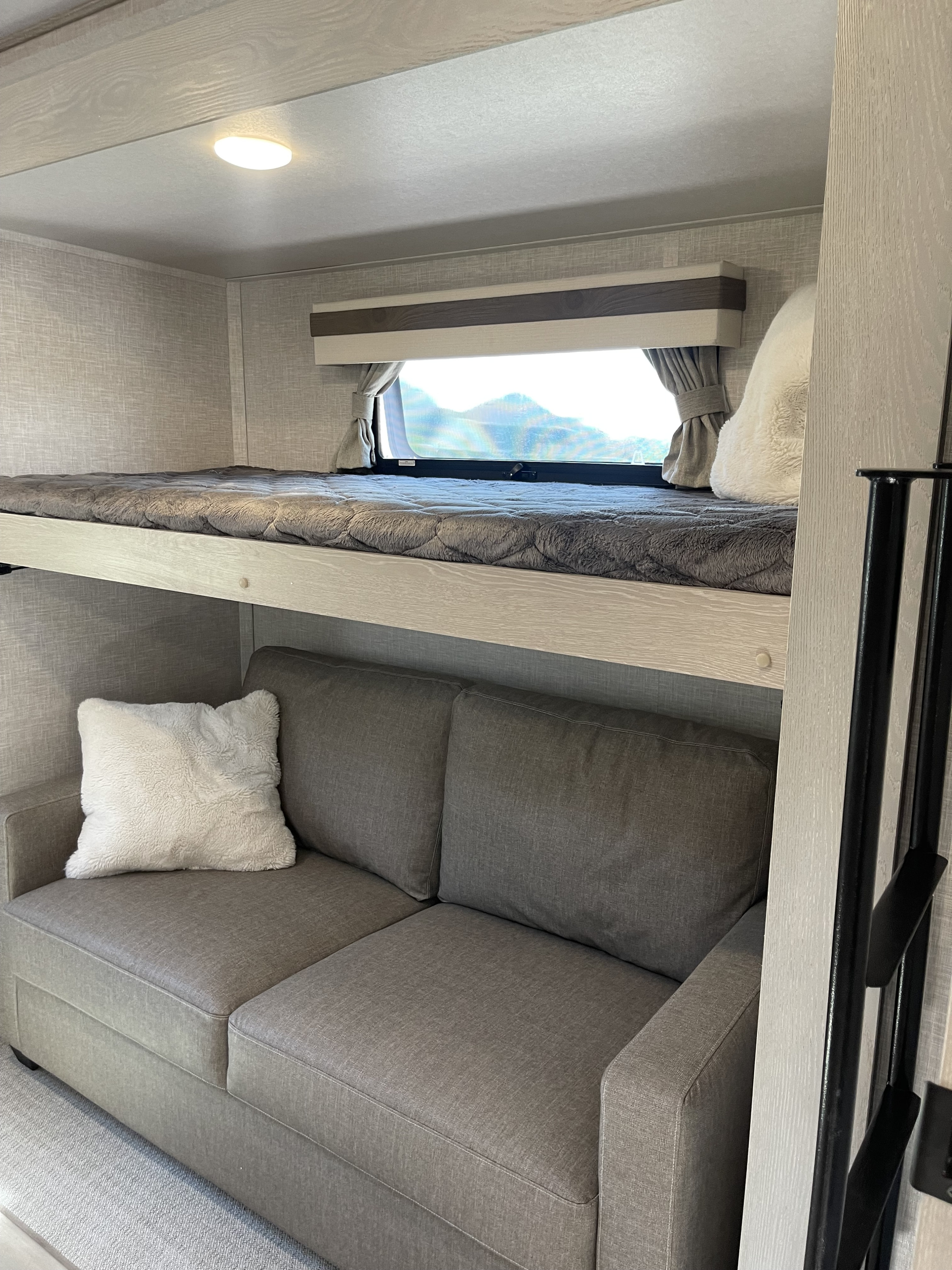 Cozy RV rental interior: upper bunk with a grey blanket, lower sofa with a white pillow, and a window framing mountain views.. Forest River Rockwood Signature 2023