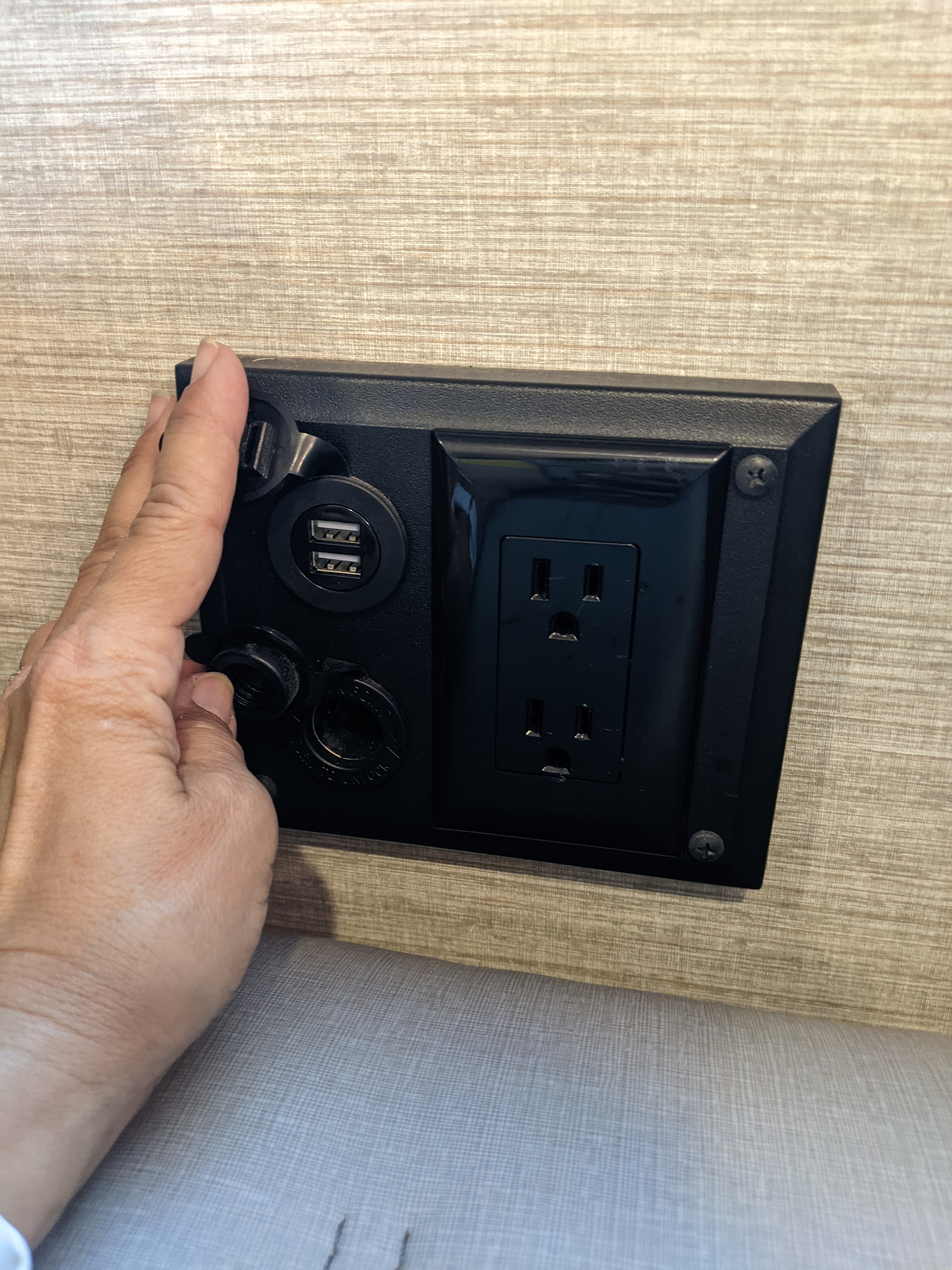 Hand accesses an RV rental's black charging station, offering USB, 12V, and standard AC power outlets for diverse device charging needs.. Winnebago Adventurer 2021