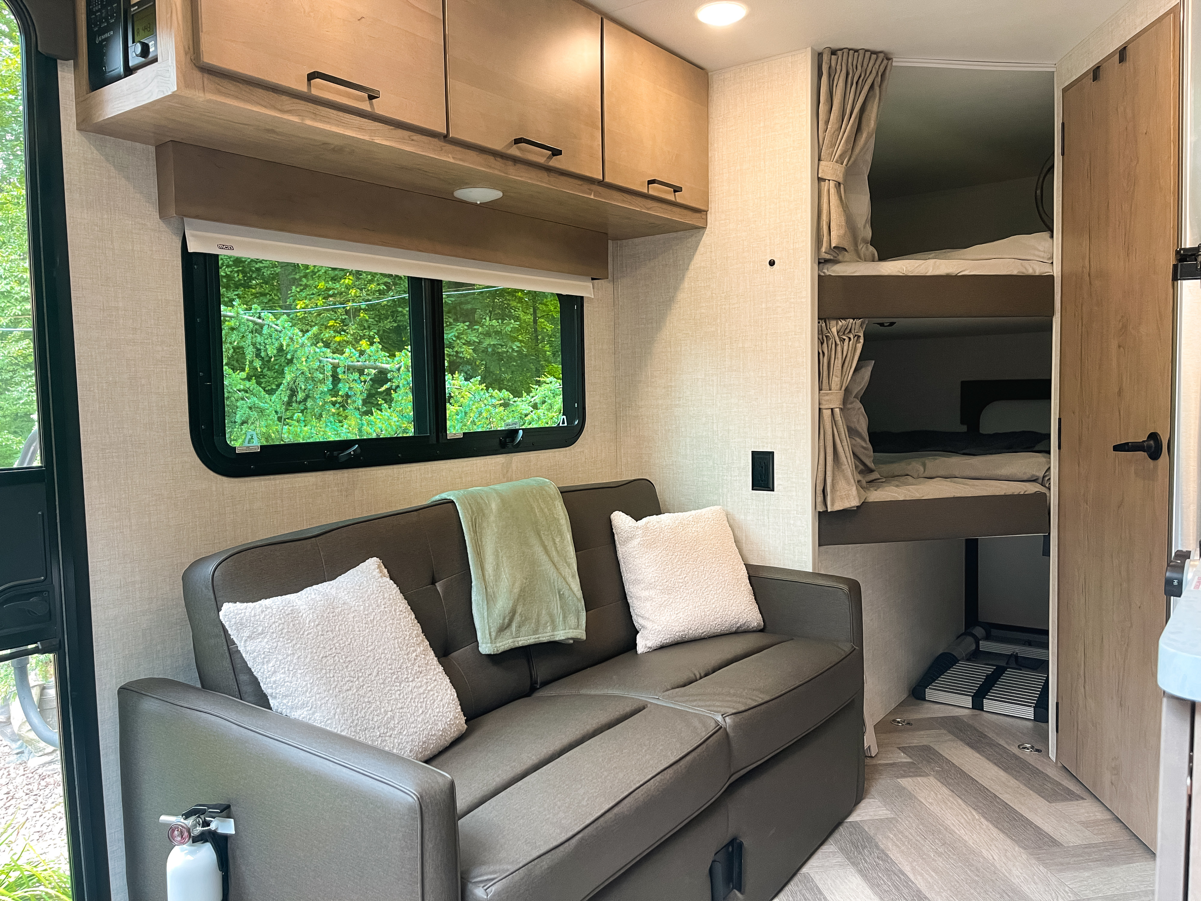 Cozy RV rental interior: a sofa with pillows, green blanket, window with trees, overhead cabinets, and bunk beds with curtains.. Ember Overland 2022