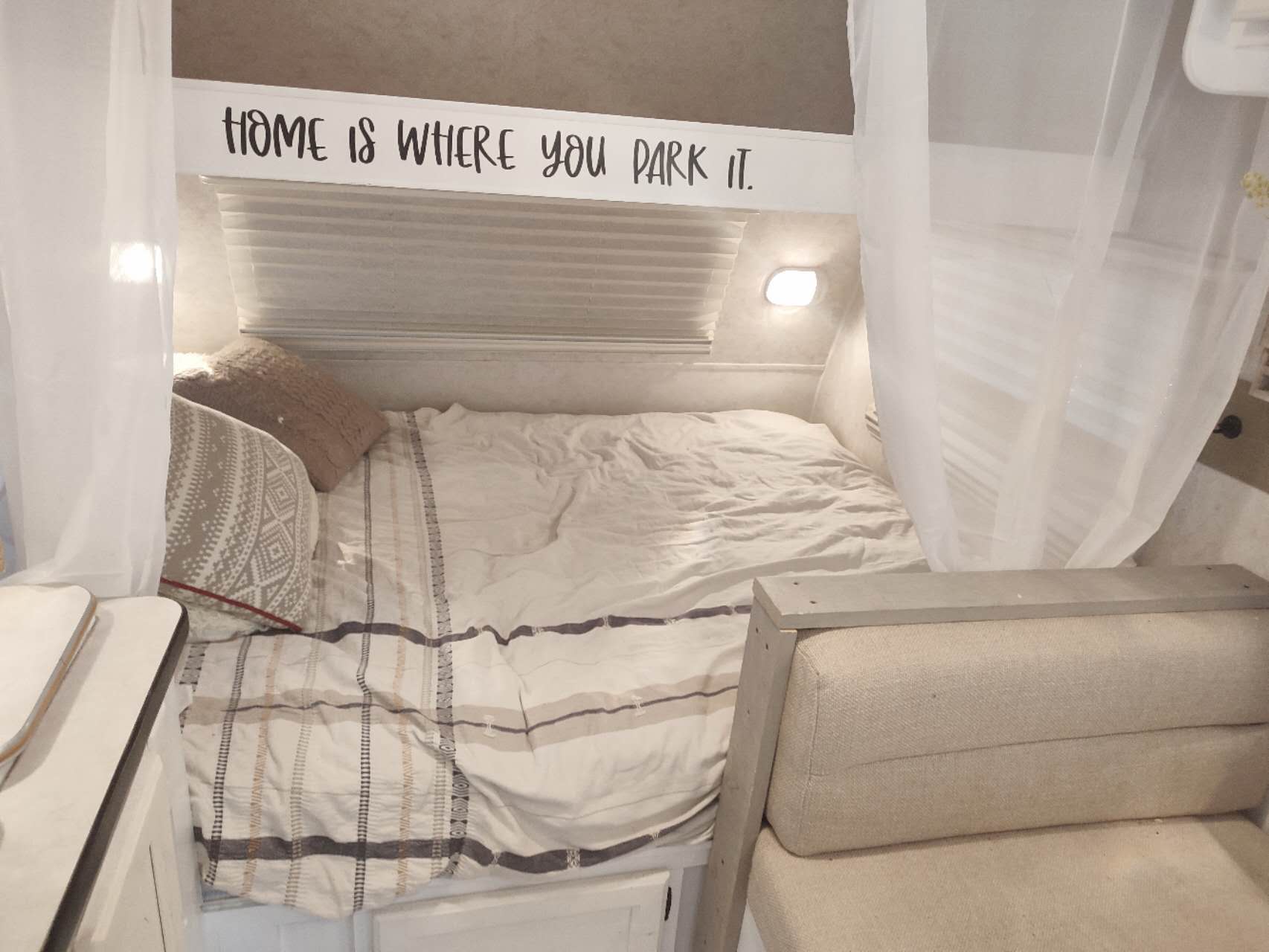 Cozy RV rental bedroom with "HOME IS WHERE YOU PARK IT" sign, bed, sheer curtains, and a seating area.. Cikira RV Classic Cruiser 2009