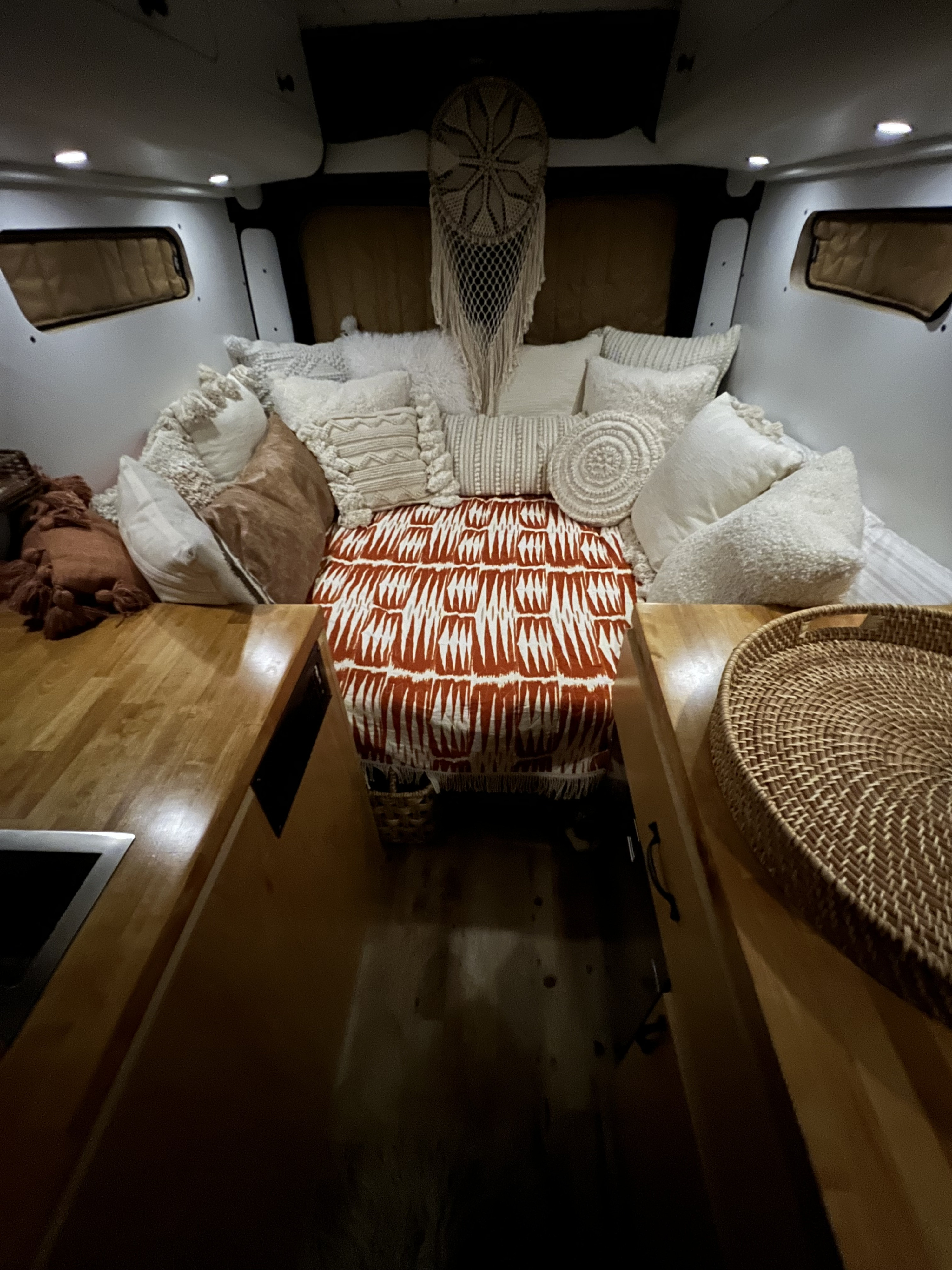 Cozy RV rental camper van bedroom with white and orange patterned bedding, macrame decor, and wooden countertops.. Dodge Ram 1500 2019