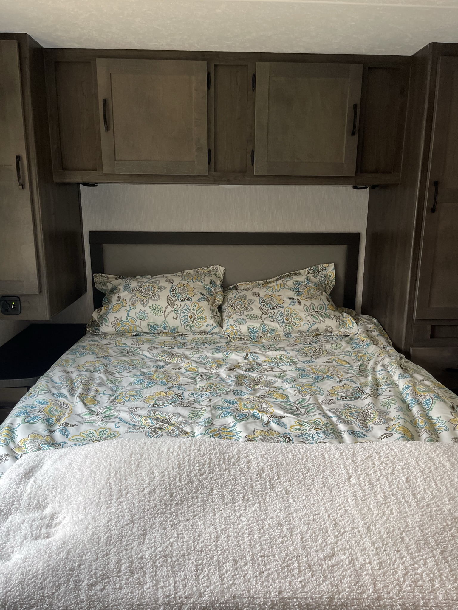 This cozy RV rental bedroom features a comfy bed with floral linens, overhead storage, and a charging port.. Coachmen Leprechaun 2023