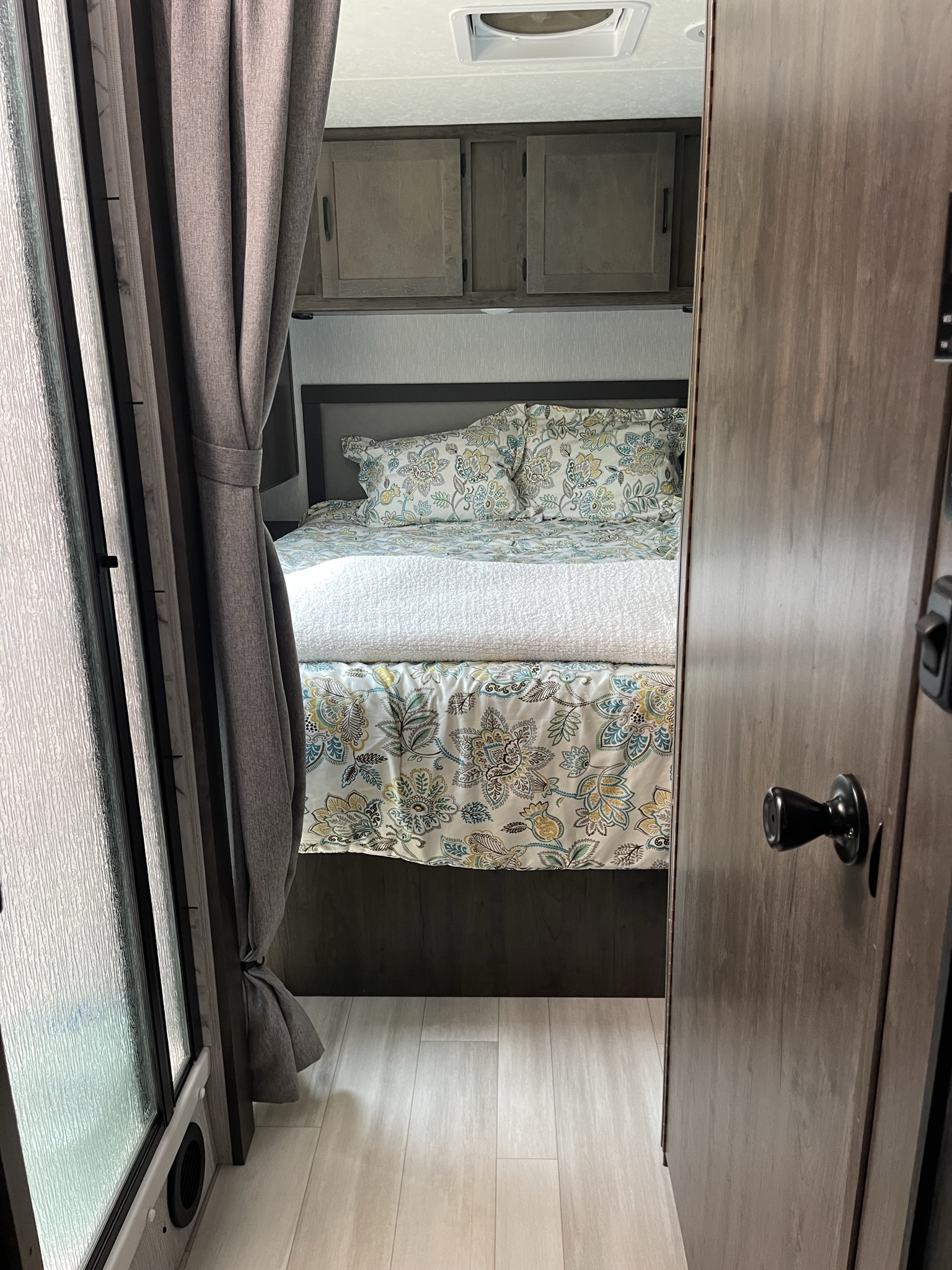 RV rental bedroom with a bed featuring floral bedding, a white blanket, overhead cabinets, and light wood-look flooring.. Coachmen Leprechaun 2023