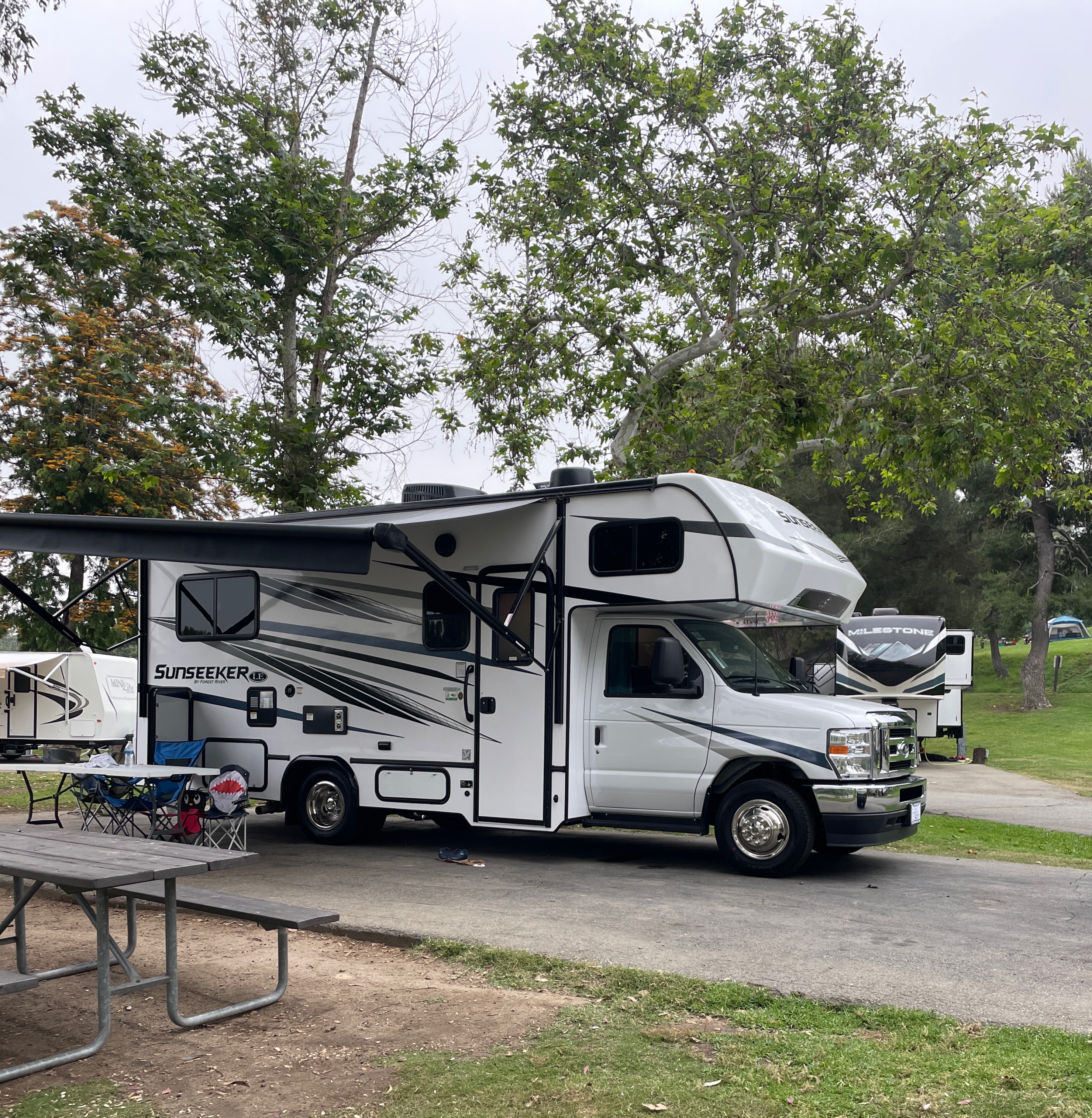 A white Sunseeker RV rental with awning extended at a campsite, with a picnic table and other RVs nearby.. Forest River Sunseeker LE 2023