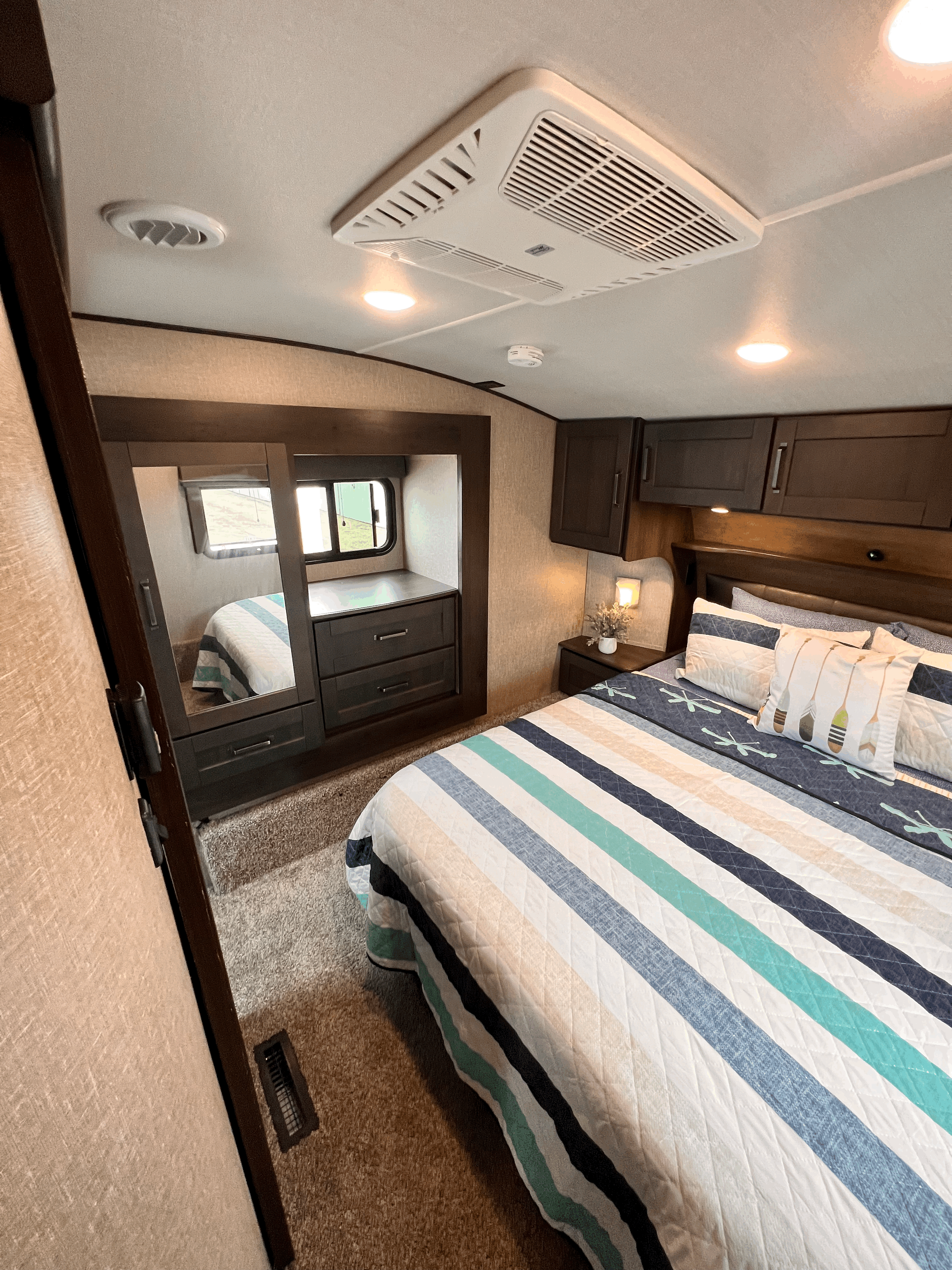 This RV rental bedroom features a comfy bed with striped quilt, mirrored closet, and overhead AC, offering convenience on your trip.. Grand Design Reflection 2023