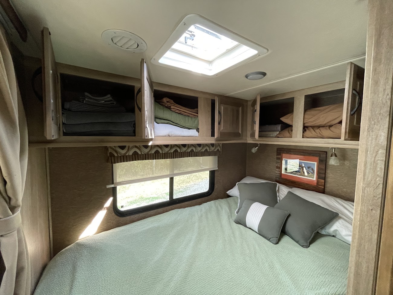Bright RV rental bedroom: bed, open overhead cabinets with linens, a window, and skylight.. Coachmen Prism 2015