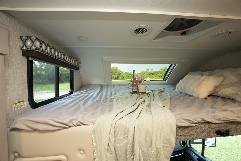 Cozy RV rental cab-over bed featuring scenic windows, soft bedding, and a drink setup. Ideal for travel and relaxation.. Thor Chateau 2023