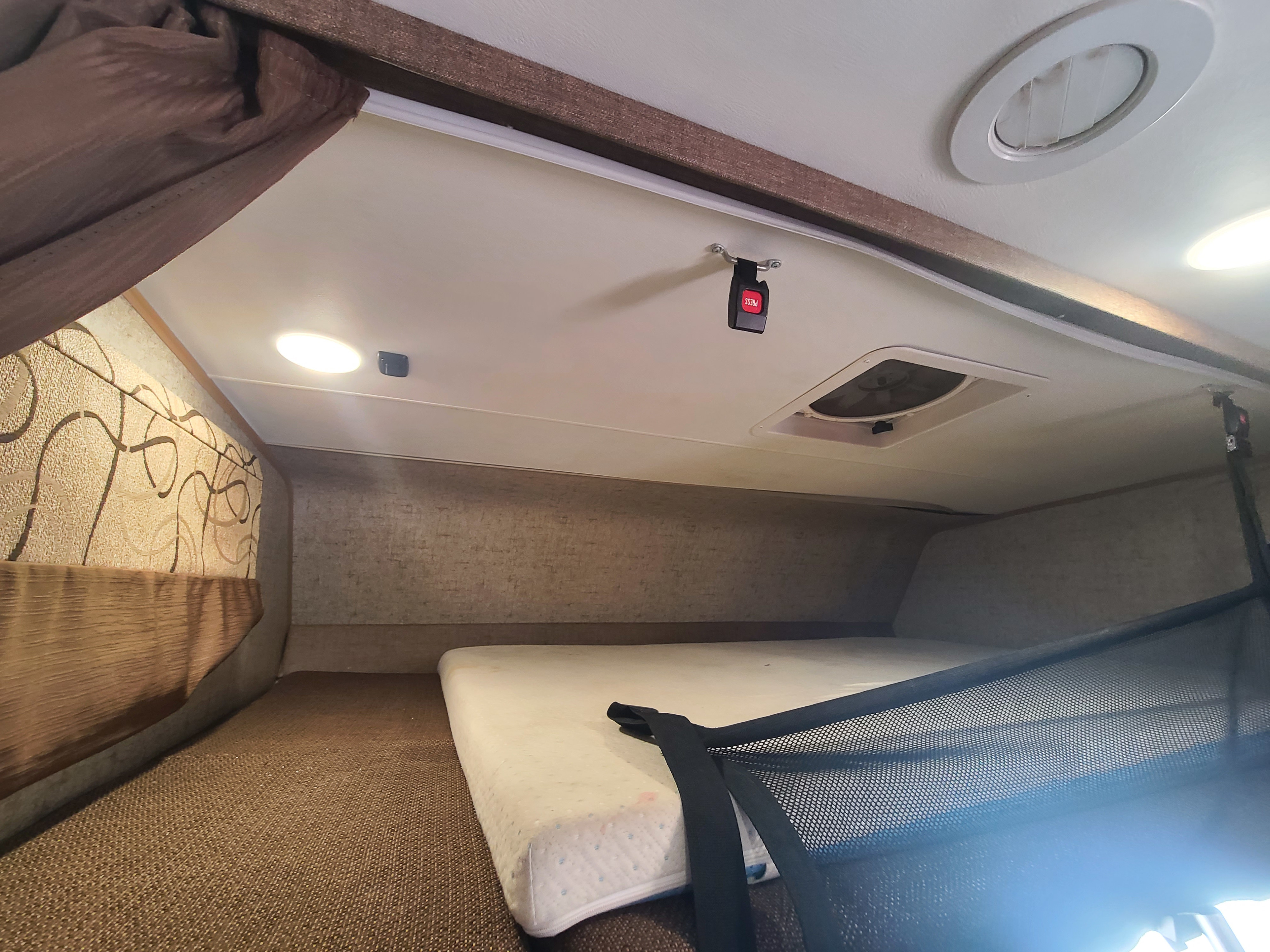 An RV rental's overhead bunk bed, showing a mattress, ceiling lights, air vent, and a mesh safety net.. Forest River Coachmen Leprechaun 2018