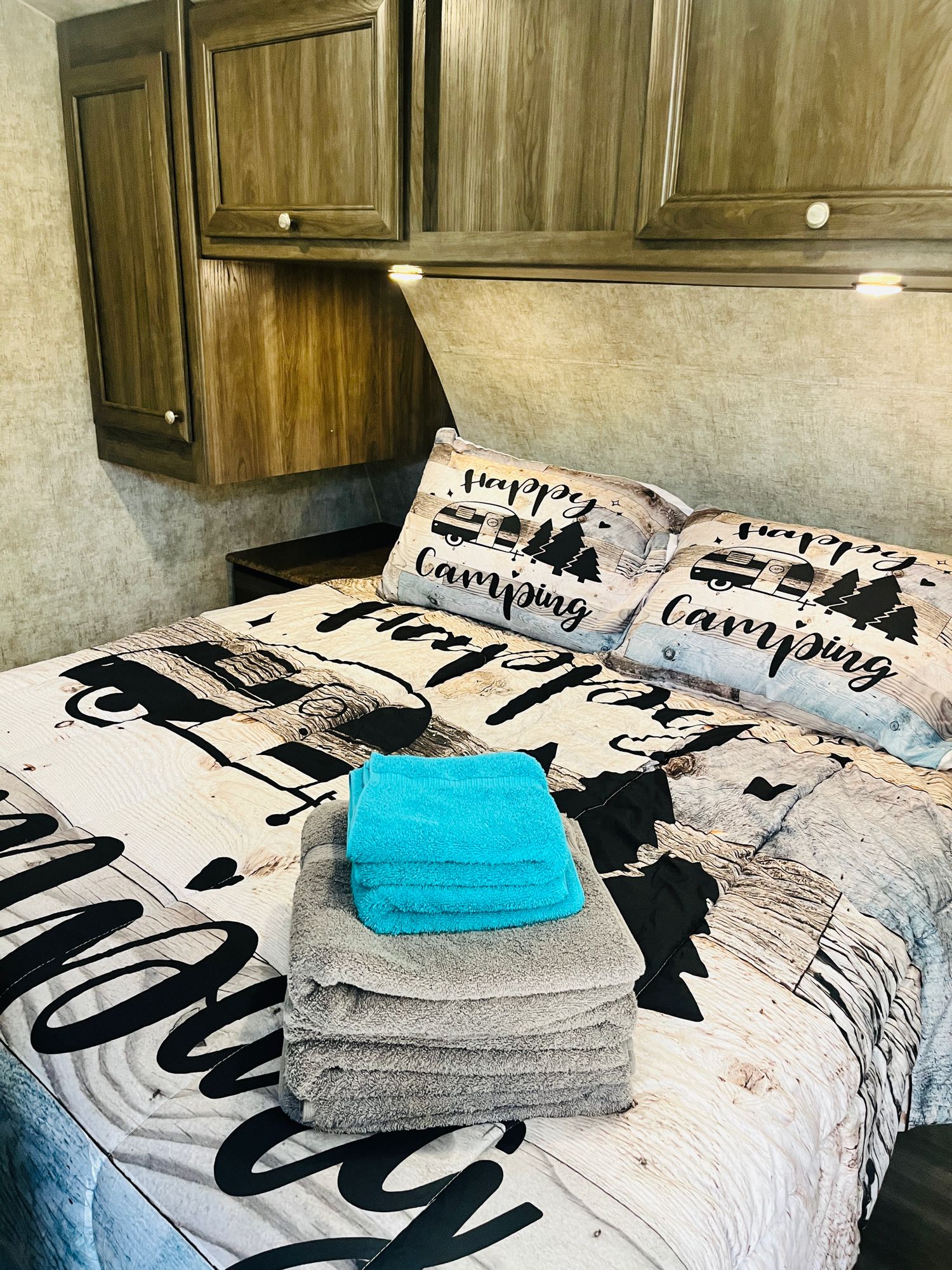 Cozy RV rental bedroom featuring "Happy Camping" bedding, wood-look cabinets, and fresh towels for a comfortable stay.. Highland Ridge RV Light 2017