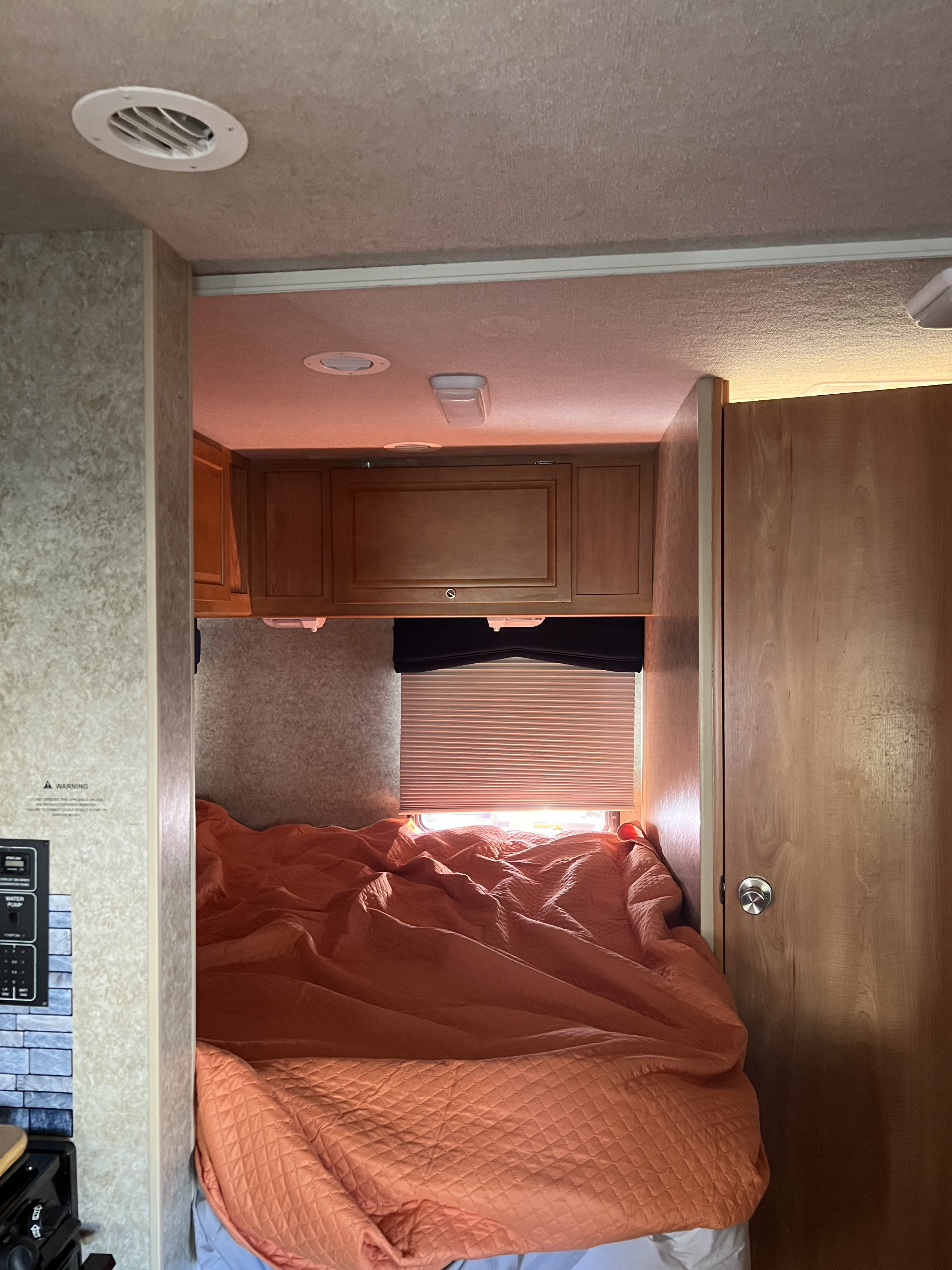 Cozy RV rental bedroom featuring a warm orange quilted comforter, overhead wooden cabinets, and a window with blinds.. Winnebago Chalet 2010