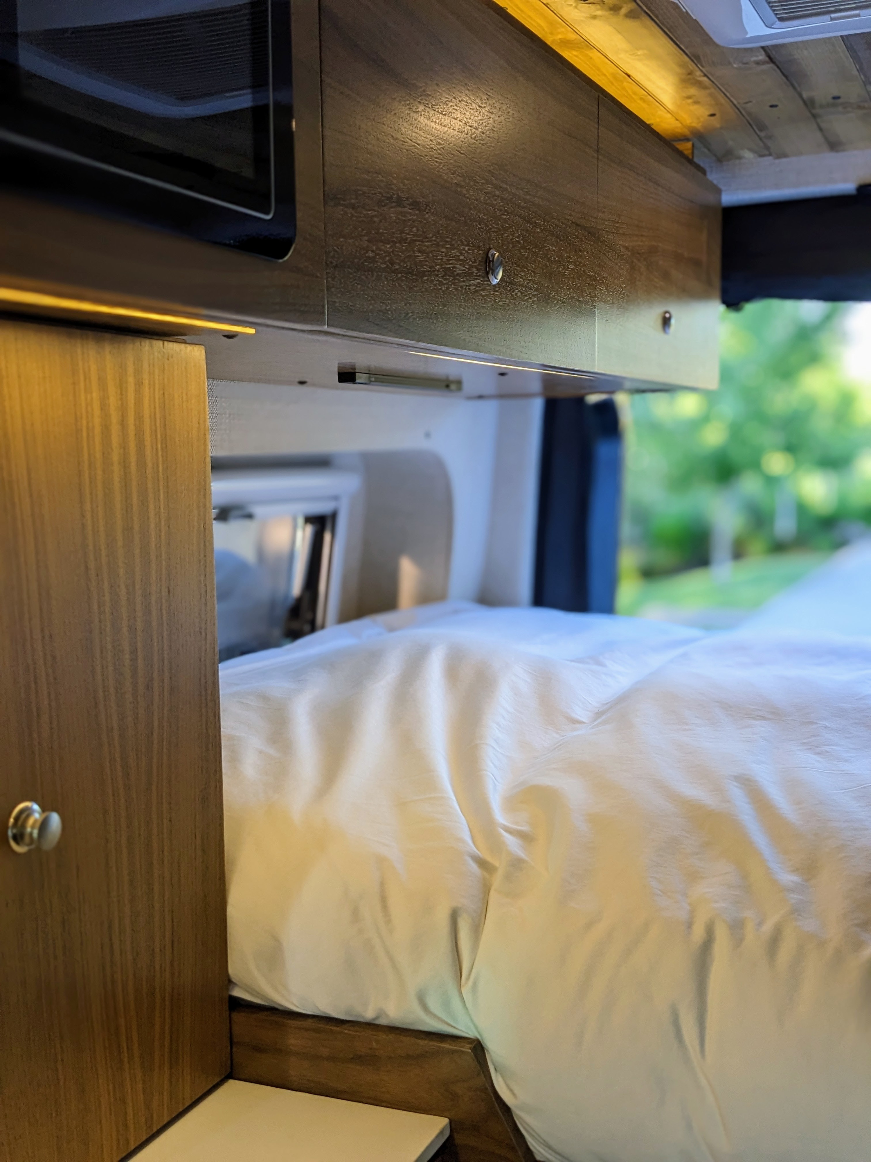 Interior of an RV rental: A warm lit sleeping area with white bedding, dark wood cabinets, and a microwave built-in above.. Mercedes-Benz Sprinter 2020