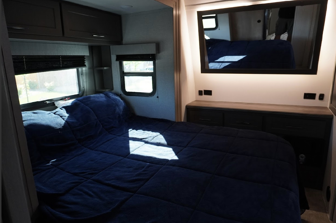 Comfortable RV rental bedroom with a blue bed, bright windows, built-in storage, and mirror. Ideal for travel.. Grand Design Momentum 2023
