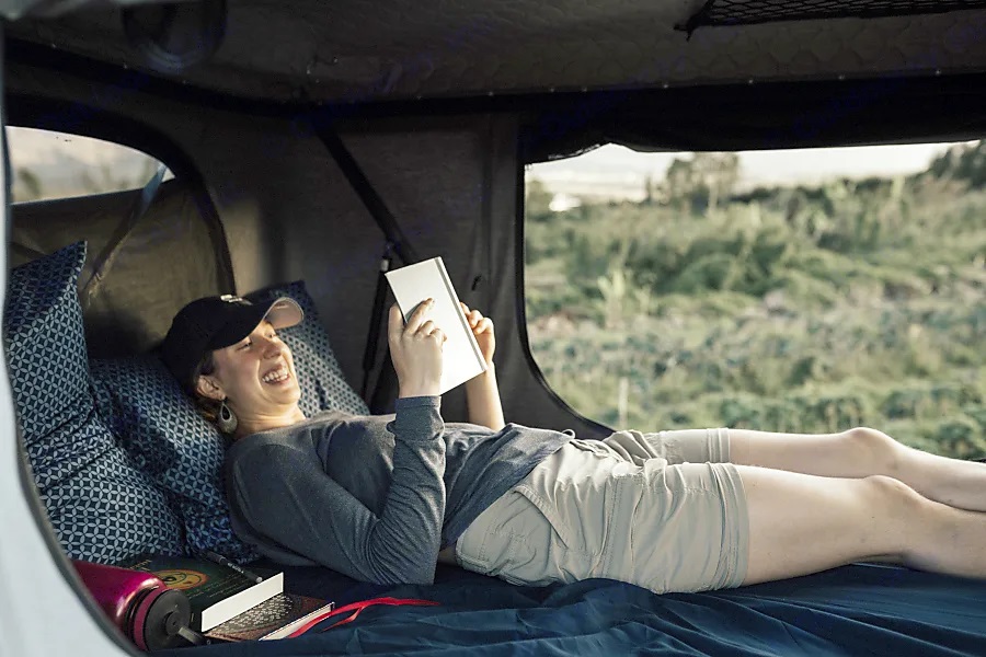 A woman laughs reading a book inside a cozy RV rental, surrounded by patterned pillows and green nature outside.. Nissan Pathfinder 2016