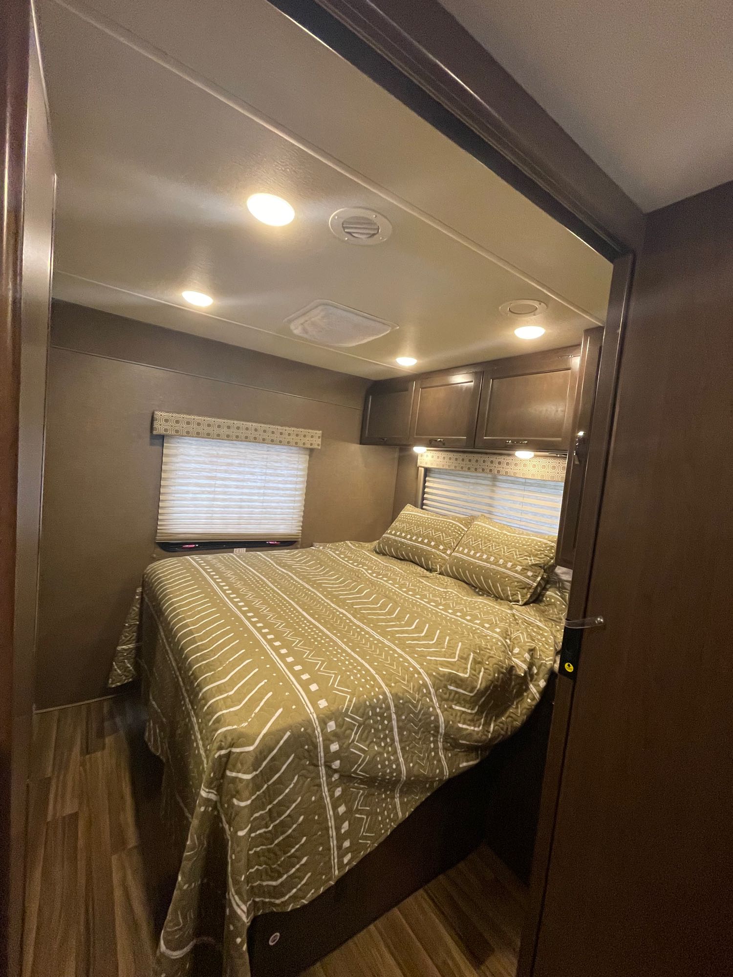 Cozy RV rental bedroom interior featuring olive patterned bedding, overhead cabinets, and window blinds.. Thor Four Winds 2019