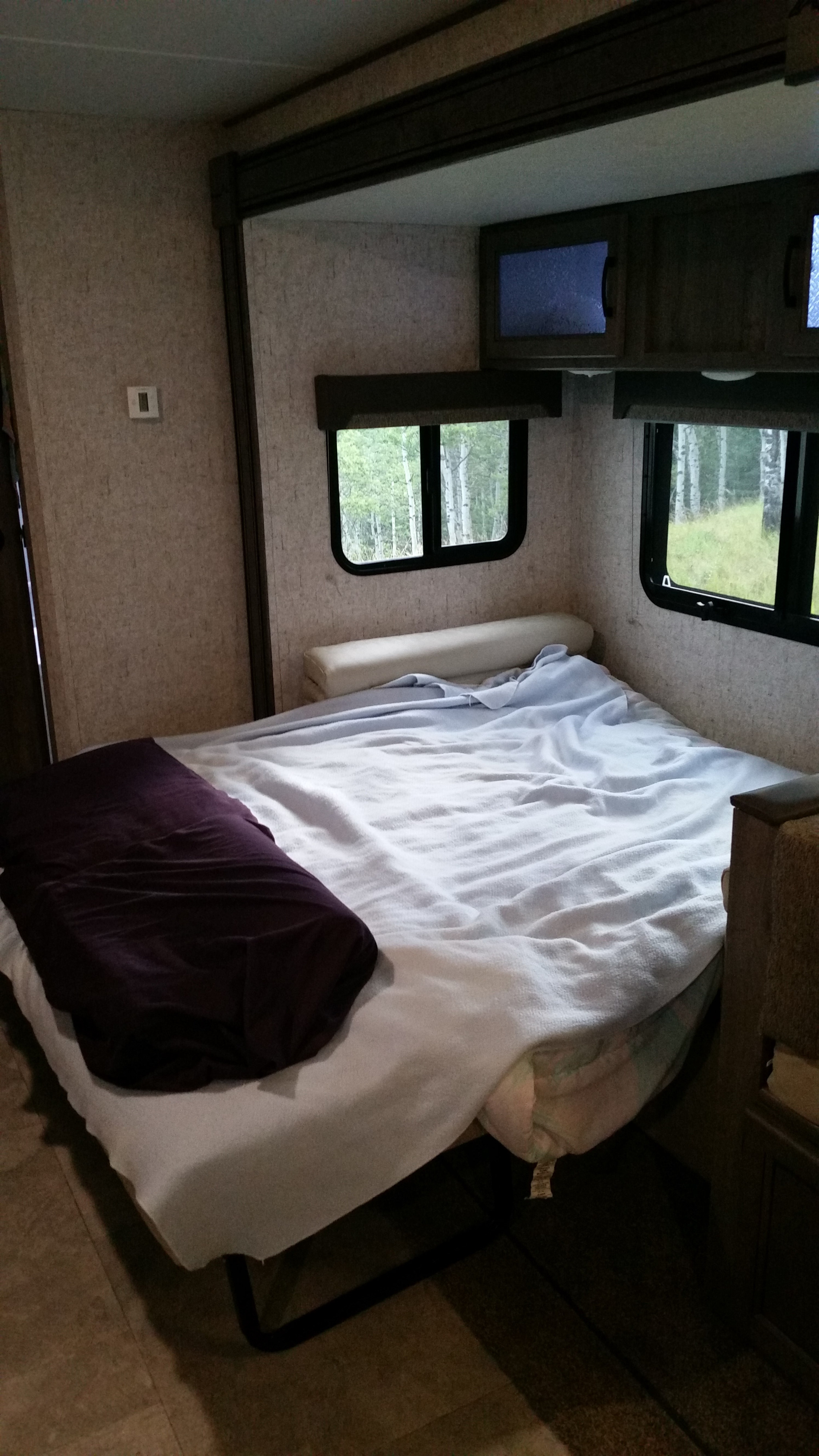 Cozy RV rental interior with a bed, windows overlooking a forest, and overhead cabinets.. Coachmen Apex 2020
