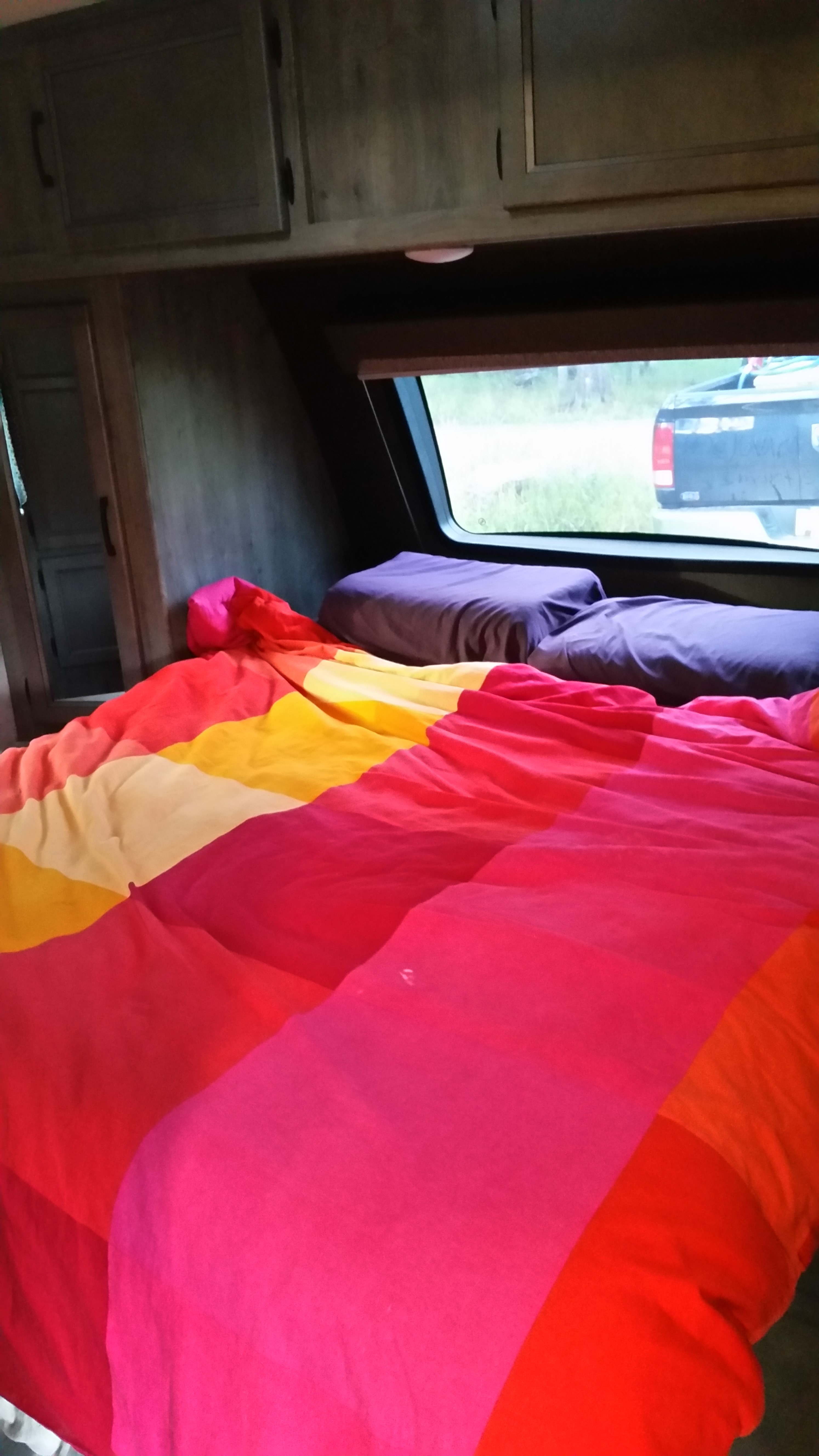 Vibrant RV rental bedroom: colorful striped bed with purple pillows, and a window overlooking a truck outside. Cozy camper interior.. Coachmen Apex 2020