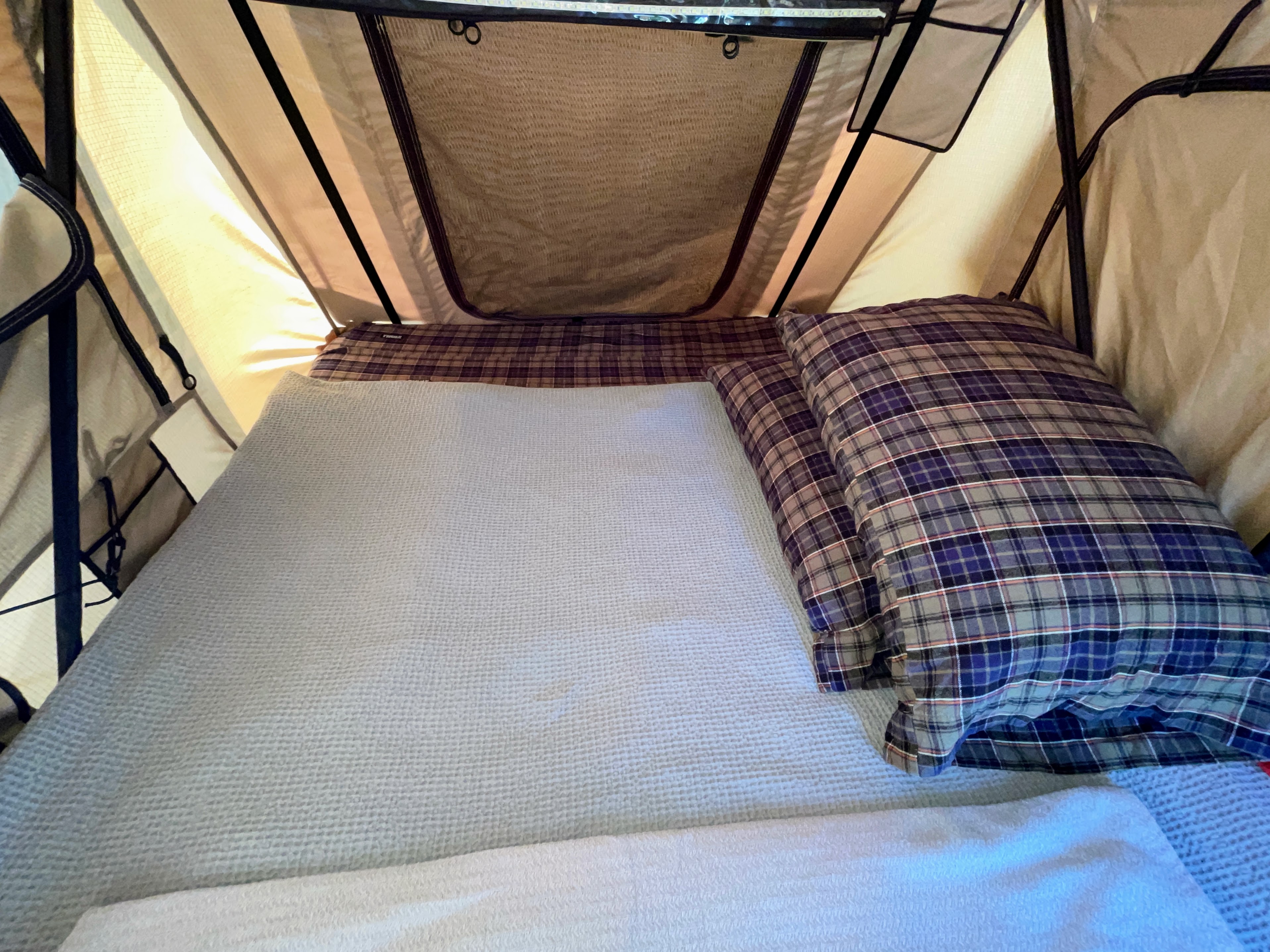 RV rental sleeping space: A cozy bed with a light grey blanket and blue plaid pillows inside a well-lit rooftop tent.. Jeep Patriot 2016