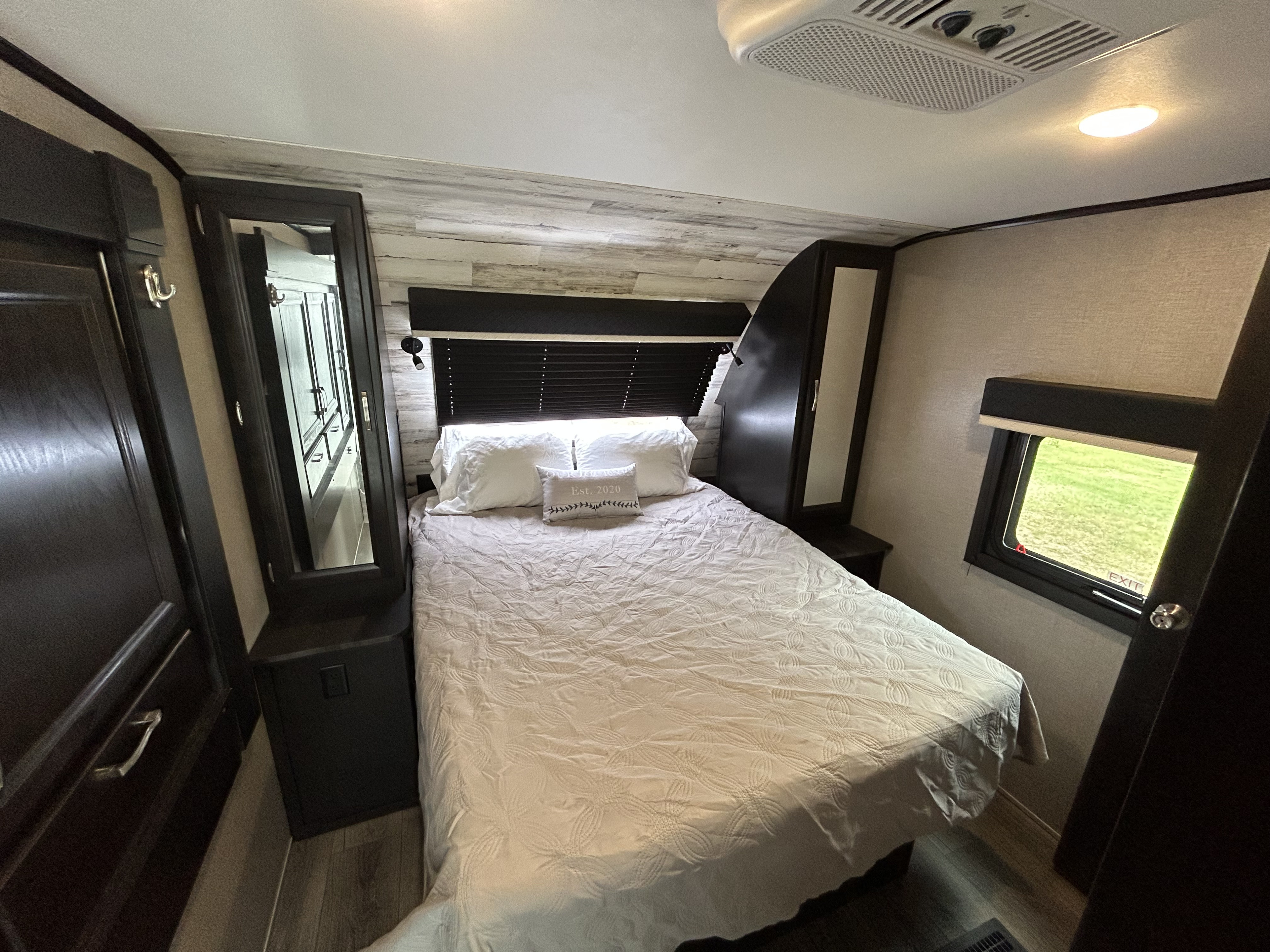 Inside a spacious RV rental bedroom featuring a full bed, dark wood cabinetry, mirror, window with green views, and ceiling AC.. Jayco White Hawk 2020