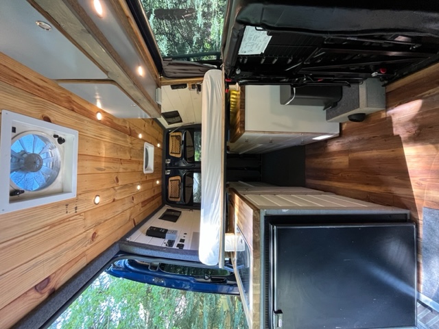 Interior view of a cozy rv rental, showcasing wood paneling, a large vent fan, lighting, and a small kitchen setup with a fridge.. Mercedes Sprinter 2016