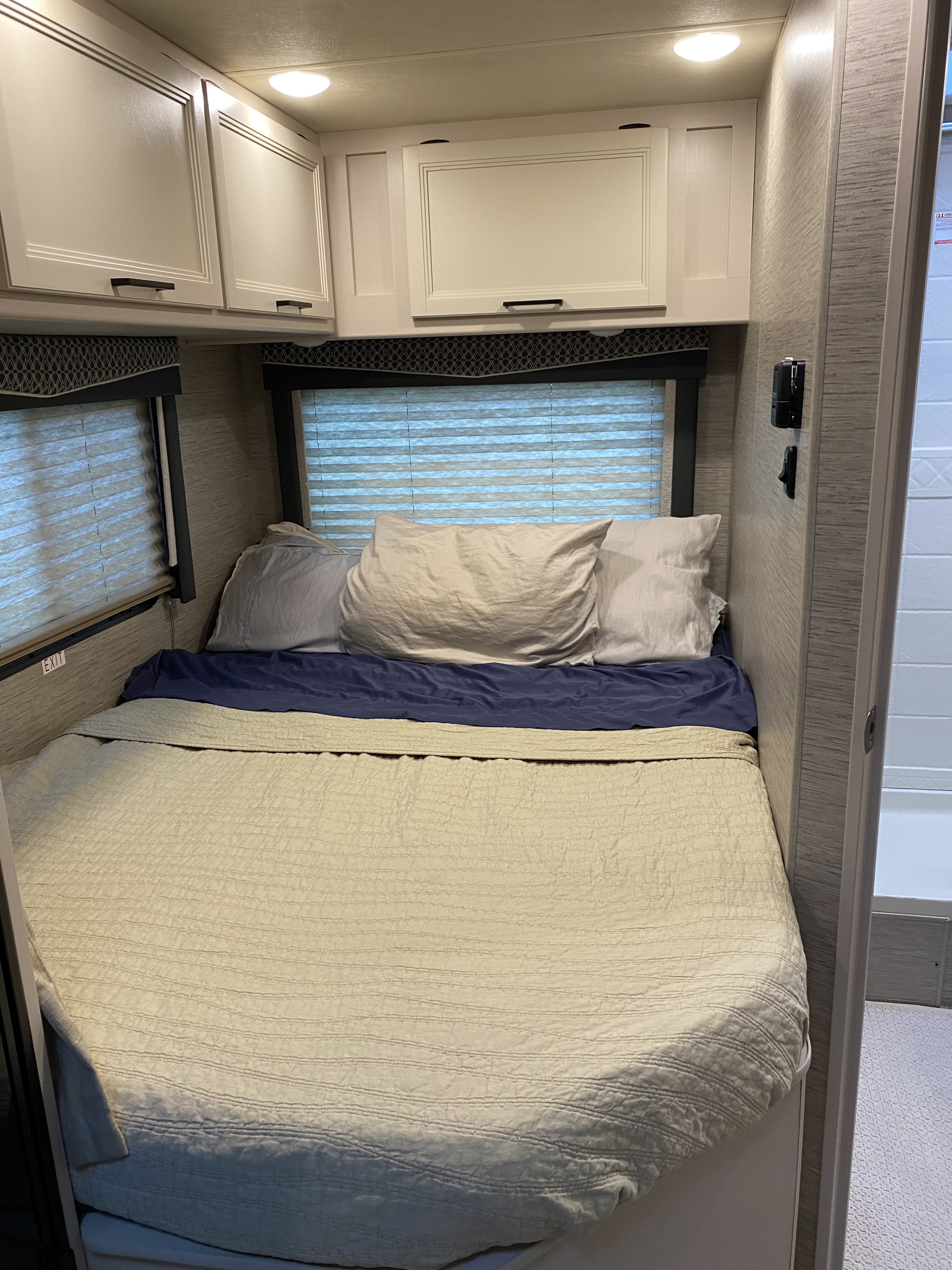 Compact RV rental bedroom featuring a bed with light-colored quilt & blue sheet, overhead cabinets, and window blinds.. Thor Four Winds 2021