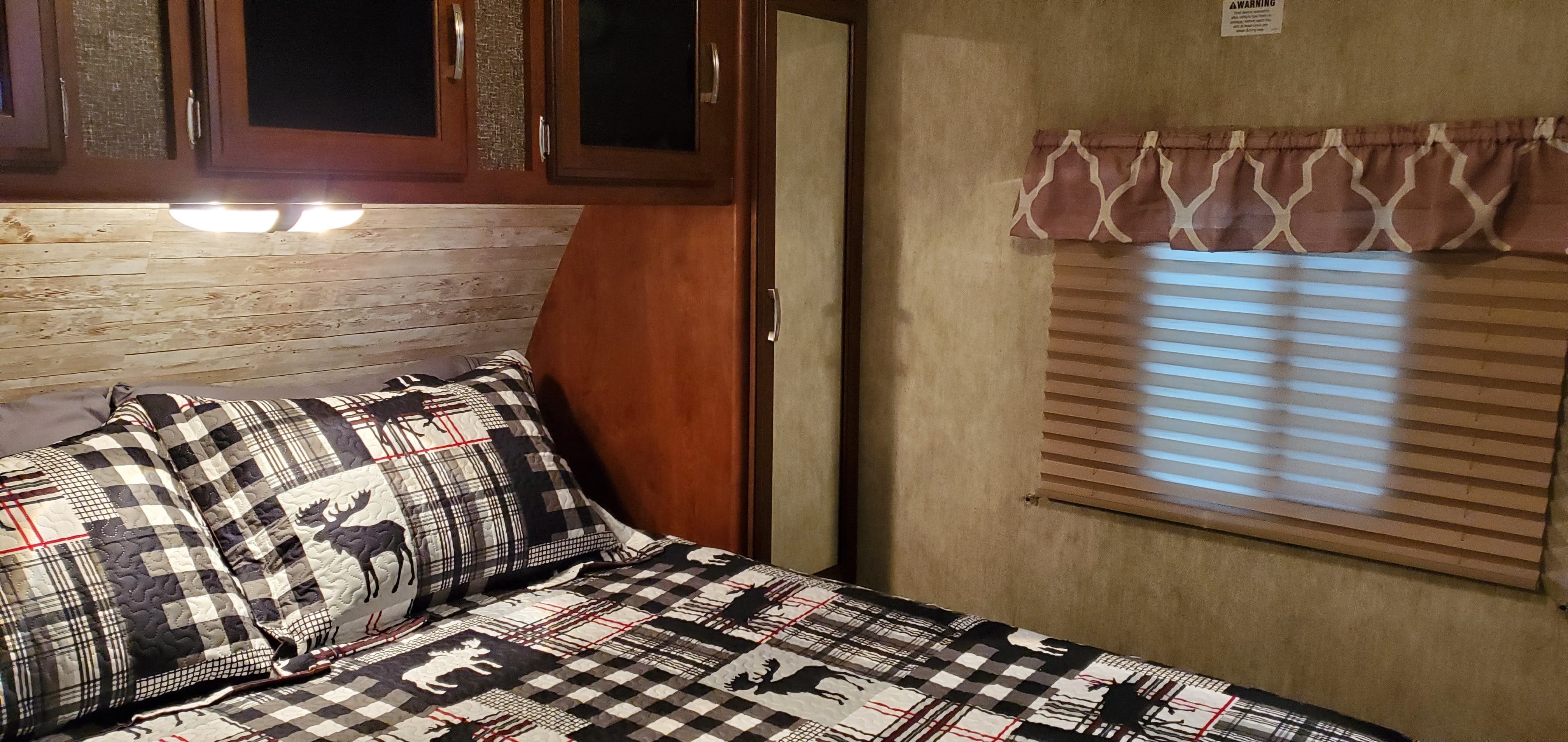 Inside an RV rental: a cozy bedroom with plaid moose bedding, wood plank headboard, and a window with blinds.. Keystone RV Passport Grand Touring 2016