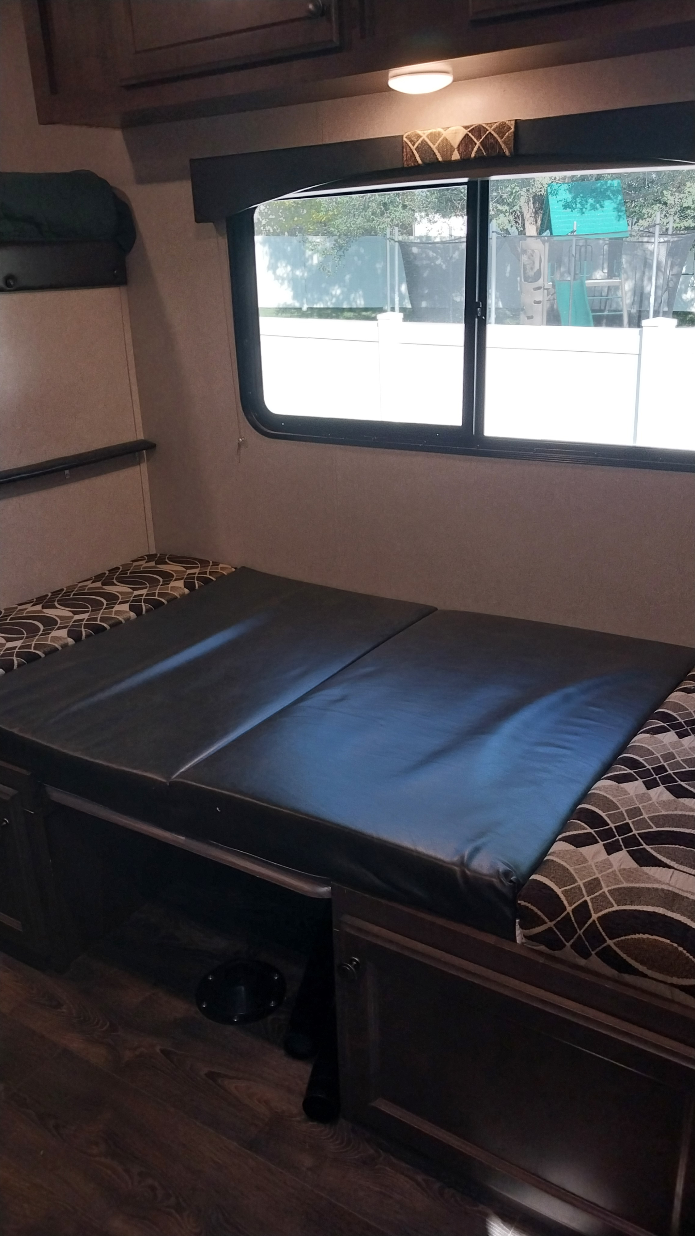 RV rental interior: A cozy dinette with black leather-like cushions and patterned backrests, converting to a bed, beneath a large window.. Sunset Park RV Sun Lite 2018