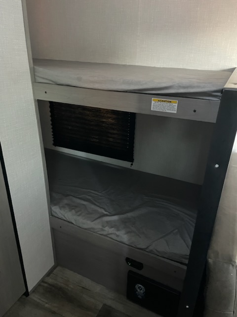 An RV rental interior showing stacked bunk beds with grey bedding, a window blind, and a lower bunk featuring a small panel.. Dutchmen Coleman Lantern LT 2023