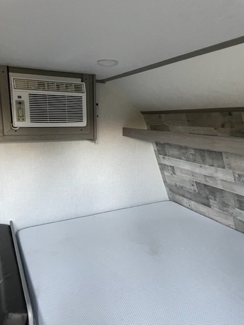Inside a cozy RV rental: compact sleeping area features a fresh mattress, wall-mounted AC unit, and stylish wood-look wall.. Dutchmen Coleman Lantern LT 2023