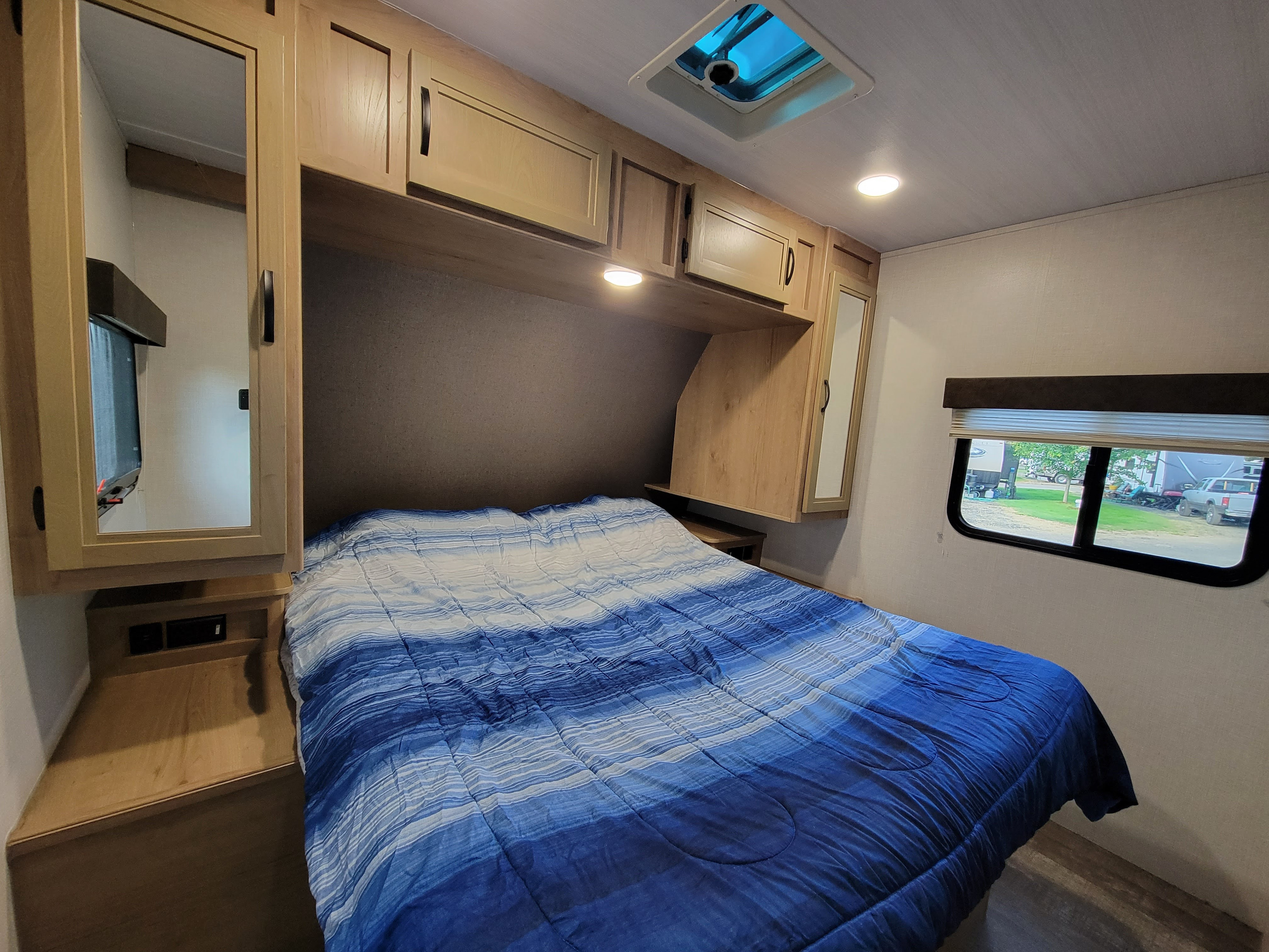 Cozy RV rental bedroom featuring a striped blue comforter, light wood cabinetry, and a side window overlooking the campsite.. Winnebago Minnie 2020