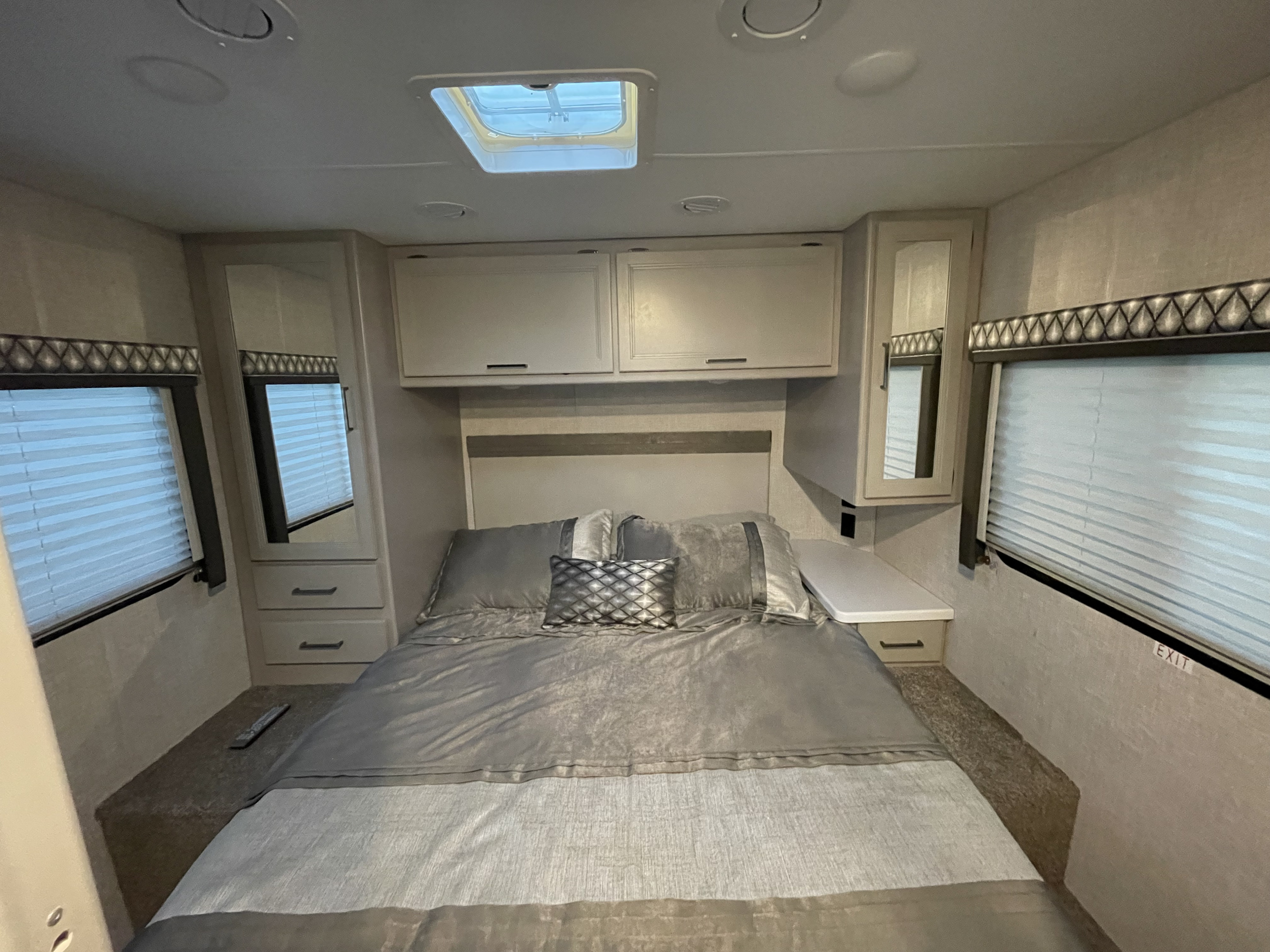 Cozy RV rental bedroom features a comfortable bed with gray linens, overhead cabinets, and large windows with blinds.. Thor Quantum LC 2022